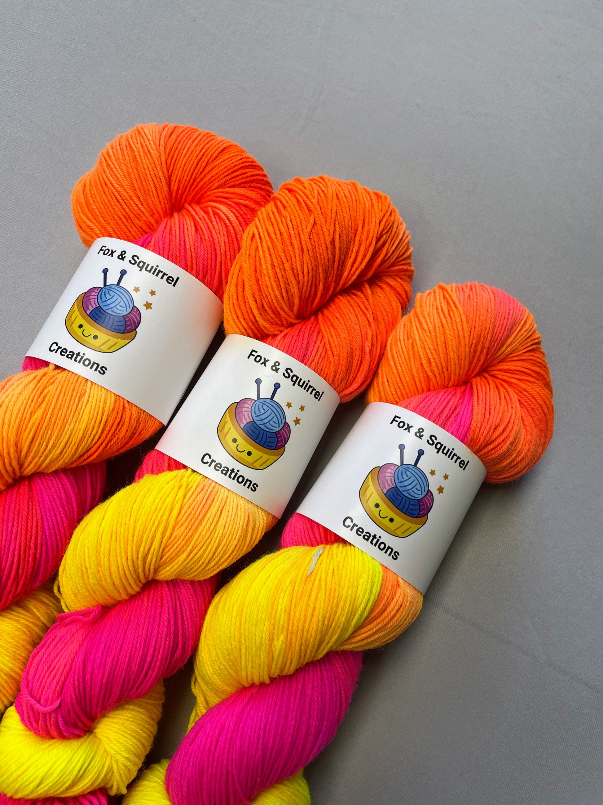 Angled shot of 100g Sock weight yarn in colourway Neon Funk. This colourway has three bands of colour in Neon Yellow, Neon Pink and Neon Orange. Photographed on a light grey background.