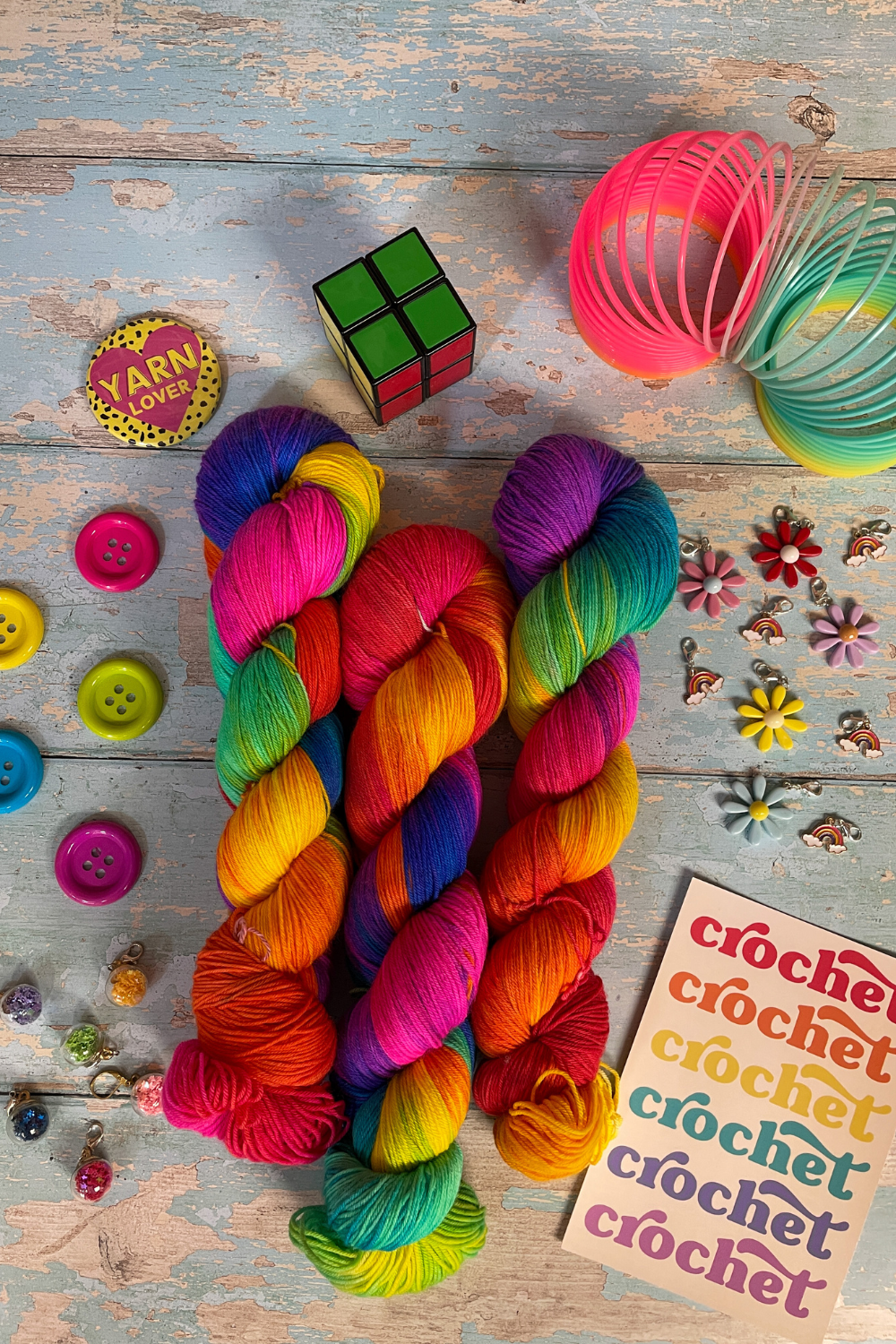 3 twisted skeins of Hand-dyed Yarn in colourway Bright Rainbow. The yarn contains 7 bright colours, sunshine, lava, ruby, apple, cyan, electric purple and neon pink. Surrounded by props, such as stitch markers and buttons on a blue, wooden background.