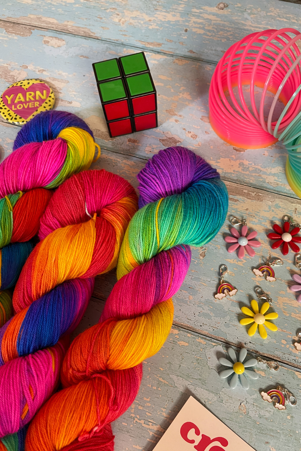 Close up photo of 3 twisted skeins of Hand-dyed Yarn in colourway Bright Rainbow. The yarn contains 7 bright colours, sunshine, lava, ruby, apple, cyan, electric purple and neon pink. Surrounded by props, such as stitch markers and rubix cube on a blue, wooden background.