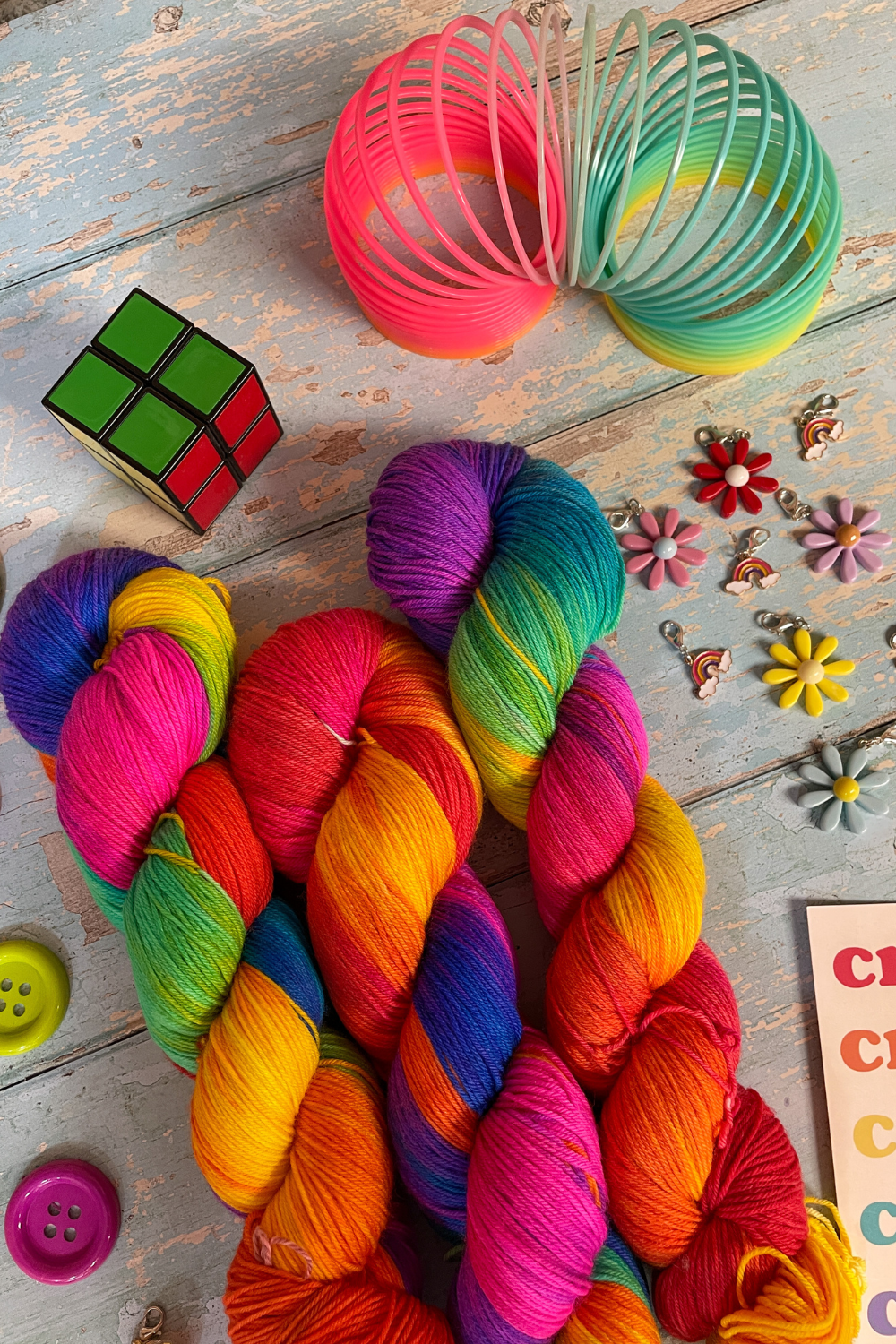 Close up photo of 3 twisted skeins of Hand-dyed Yarn in colourway Bright Rainbow. The yarn contains 7 bright colours, sunshine, lava, ruby, apple, cyan, electric purple and neon pink. Surrounded by props, such as stitch markers and slinky on a blue, wooden background.
