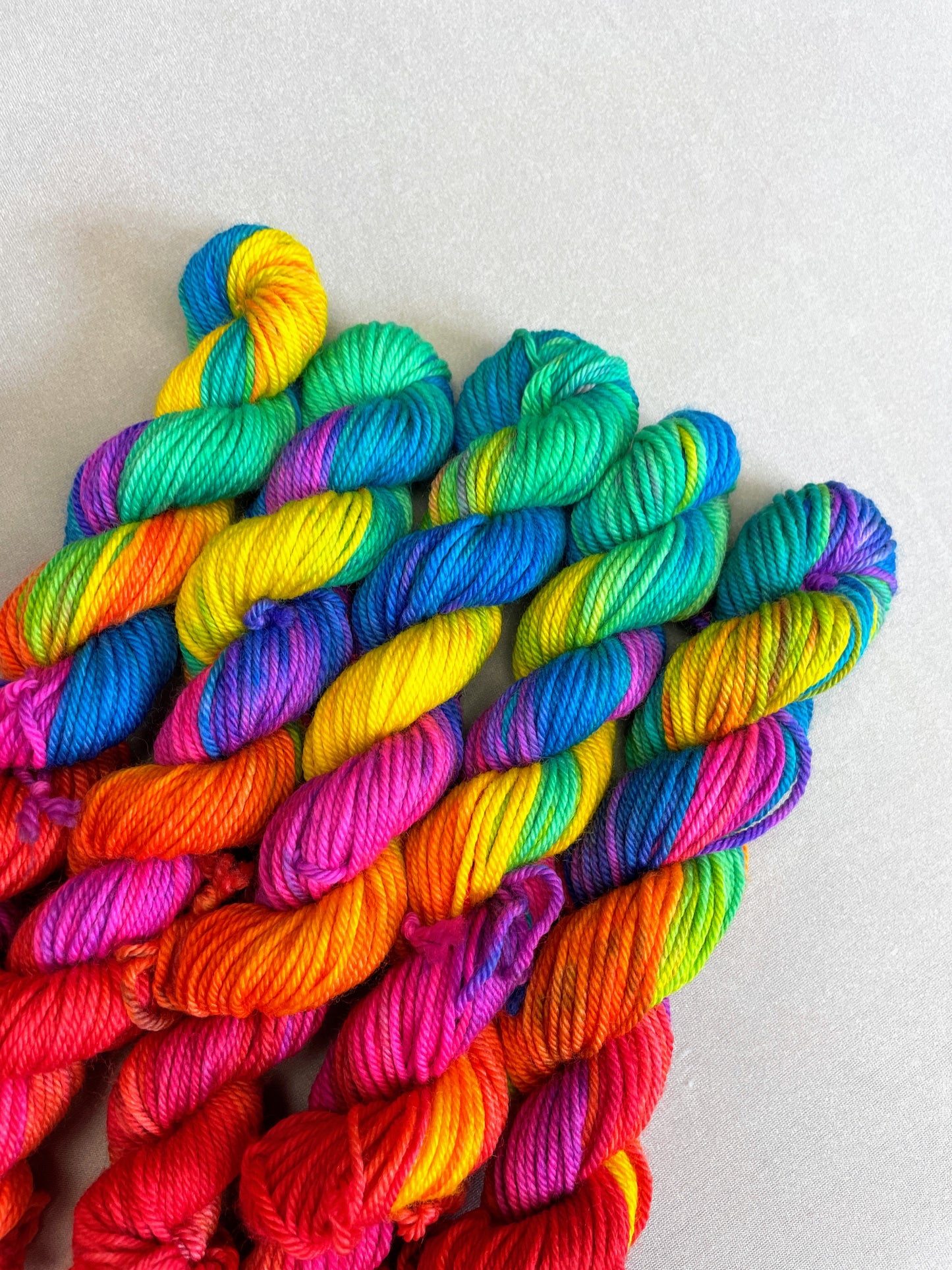 5 x 20g Hand-dyed Yarn mini skeins in colourway Bright Rainbow. The image shows a close up view of the twisted skeins on our DK weight Yarn Base. The yarn contains 7 bright colours, sunshine, lava, ruby, apple, cyan, electric purple and neon pink and photographed on a white background.