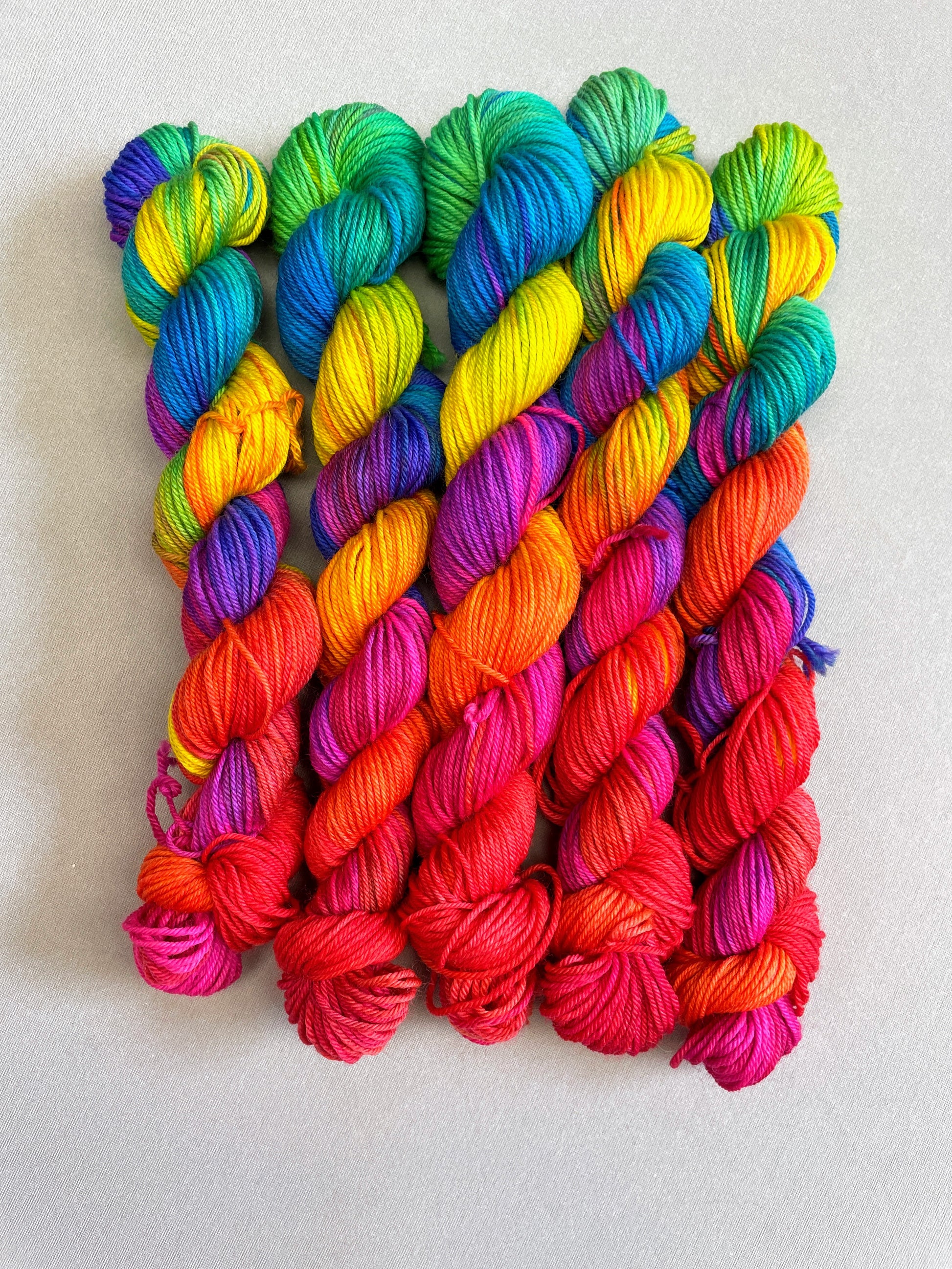 5 x 20g Hand-dyed Yarn mini skeins in colourway Bright Rainbow on our Sock/4ply Yarn base. The image shows the full skeins, twisted up and is on a Sock/4ply weight Yarn Base. The yarn contains 7 bright colours, sunshine, lava, ruby, apple, cyan, electric purple and neon pink and photographed on white background.