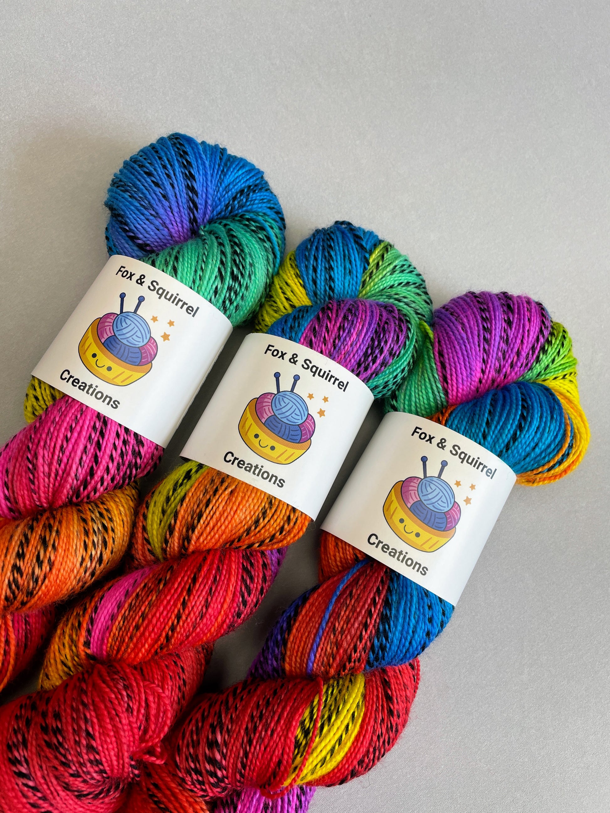 Close up shot of 3 skeins of Hand-dyed Yarn in colourway Bright Rainbow on our Zebra Sock yarn base. The yarn contains 7 bright colours, sunshine, lava, ruby, apple, cyan, electric purple and neon pink and is photographed on a white background.