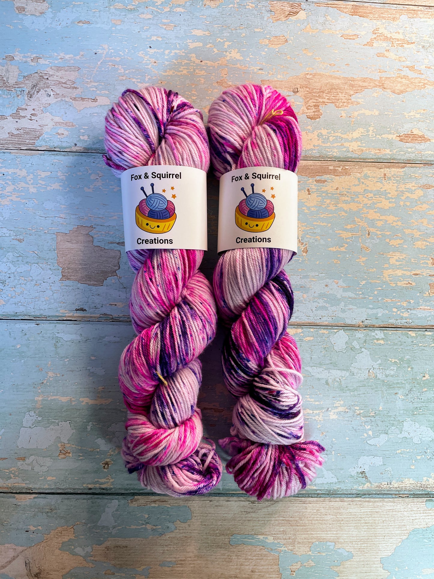 Full shot of 100g DK weight yarn in colourway Berry Sprinkles. An undyed yarn with layers of speckles in Pink, Neon Pink and Purple. Photographed on a blue wooden background.