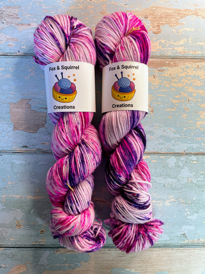 Zoomed in shot of 100g DK weight yarn in colourway Berry Sprinkles. An undyed yarn with layers of speckles in Pink, Neon Pink and Purple. Photographed on a blue wooden background.