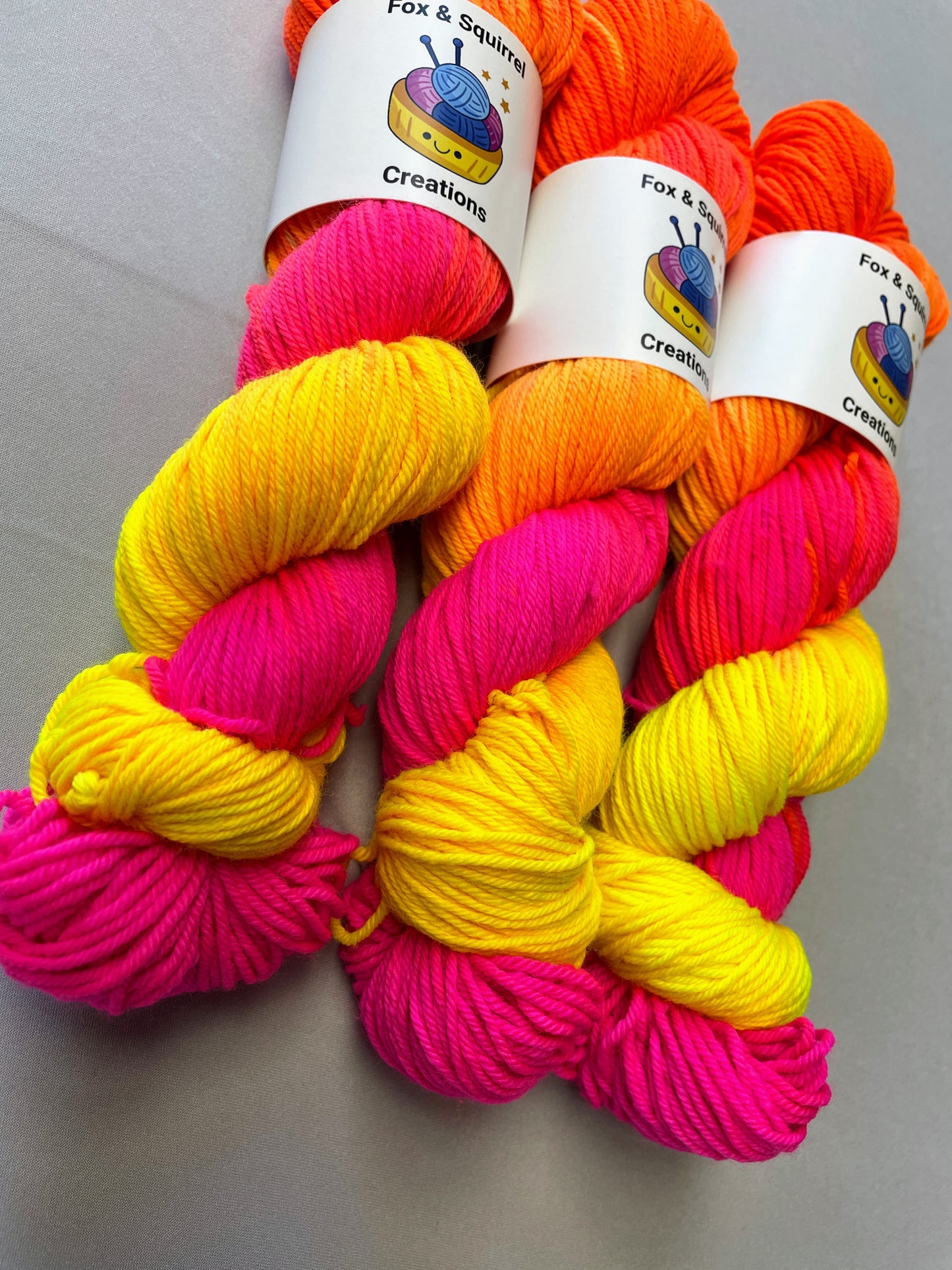 Close up shot of 100g DK weight yarn in colourway Neon Funk. This colourway has three bands of colour in Neon Yellow, Neon Pink and Neon Orange. Photographed on a light grey background. 