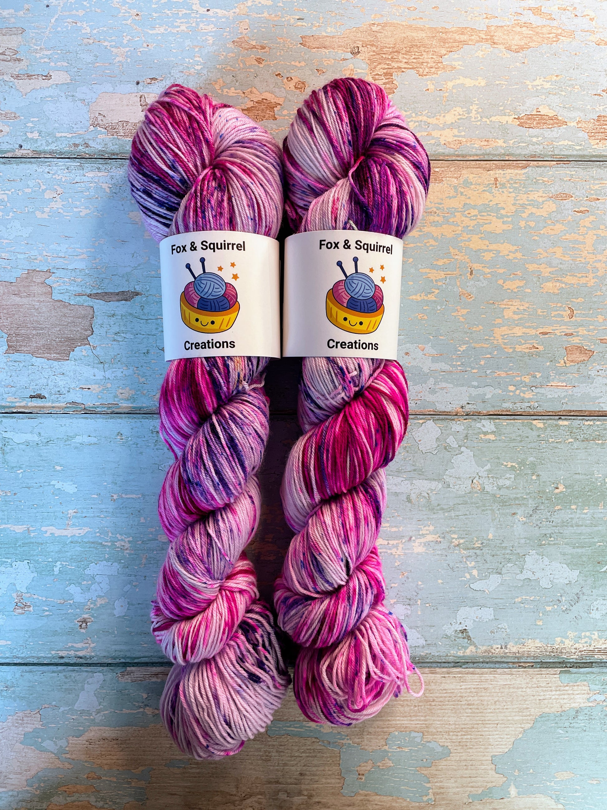 Full shot of 100g Sock weight yarn in colourway Berry Sprinkles. An undyed yarn with layers of speckles in Pink, Neon Pink and Purple. Photographed on a blue wooden background.