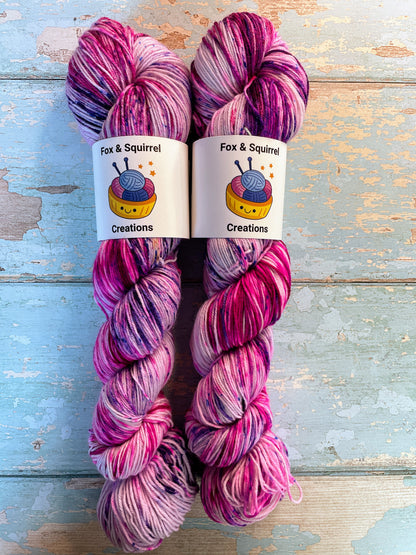 Zoomed in shot of 100g Sock weight yarn in colourway Berry Sprinkles. An undyed yarn with layers of speckles in Pink, Neon Pink and Purple. Photographed on a blue wooden background.