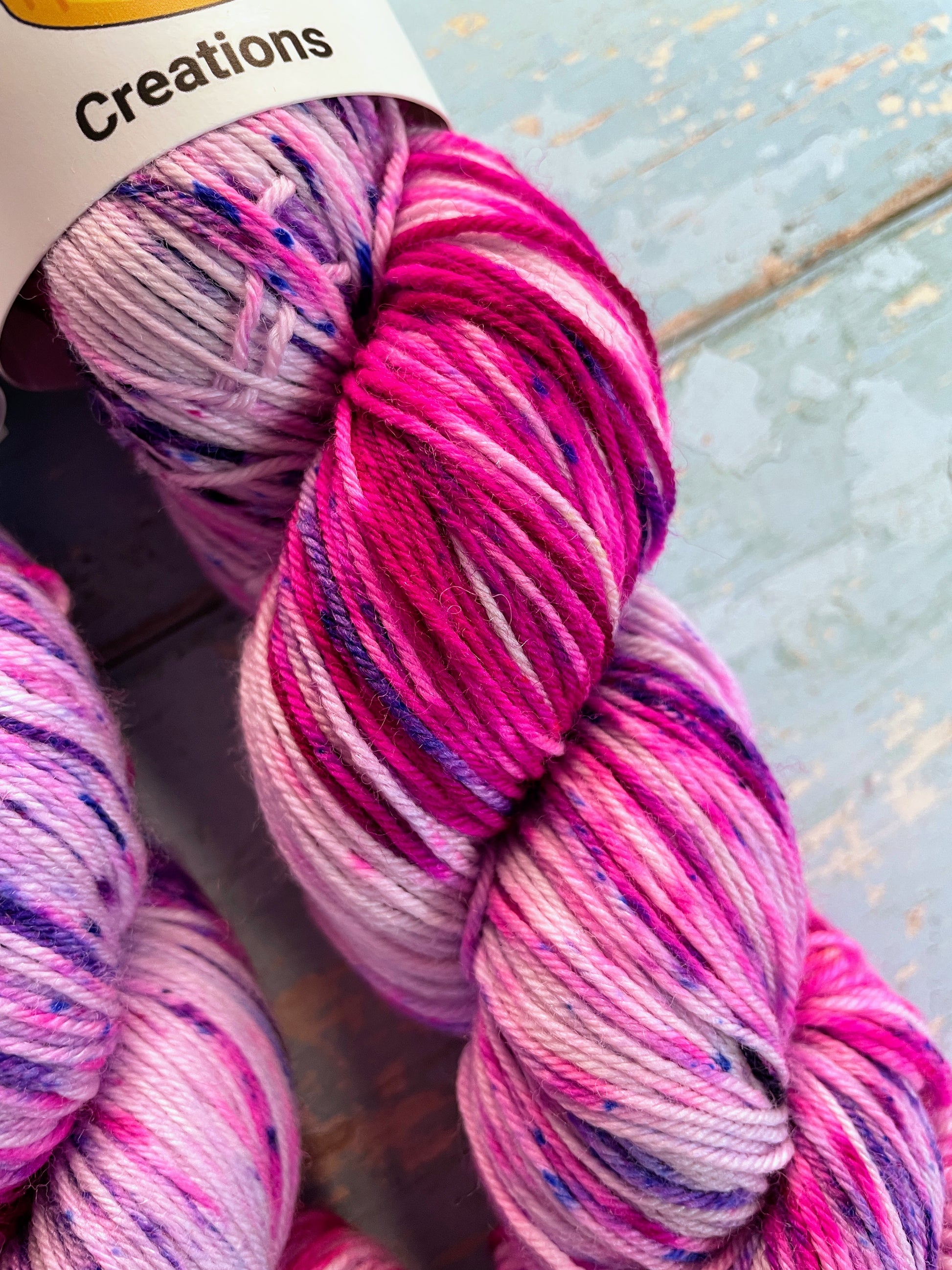 Close up shot of 100g Sock weight yarn in colourway Berry Sprinkles. An undyed yarn with layers of speckles in Pink, Neon Pink and Purple. Photographed on a blue wooden background.