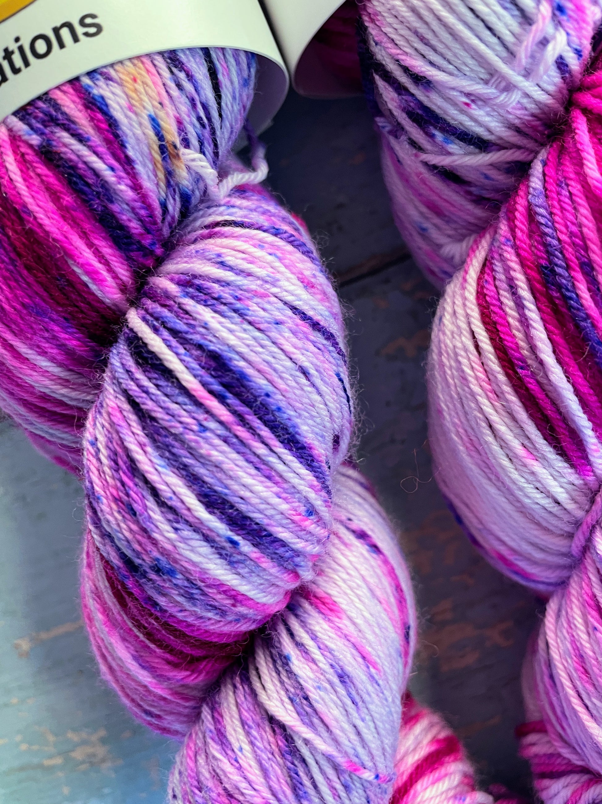 Close up of speckles of 100g Sock weight yarn in colourway Berry Sprinkles. An undyed yarn with layers of speckles in Pink, Neon Pink and Purple. Photographed on a blue wooden background.