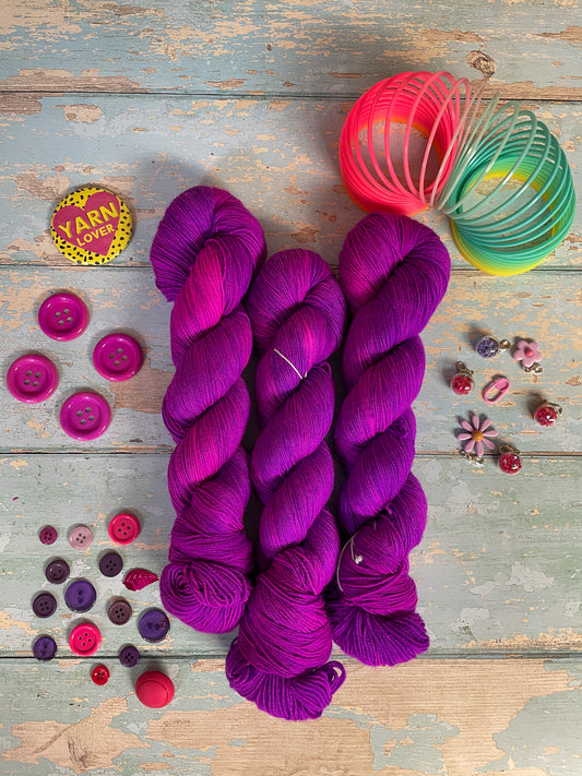 Full shot of 100g sock weight yarn in semi-solid colourway Electric Purple. This colourway is an intense purple that breaks to neon pink. Photographed on blue wooden background surrounded by props such as buttons and stitch markers.