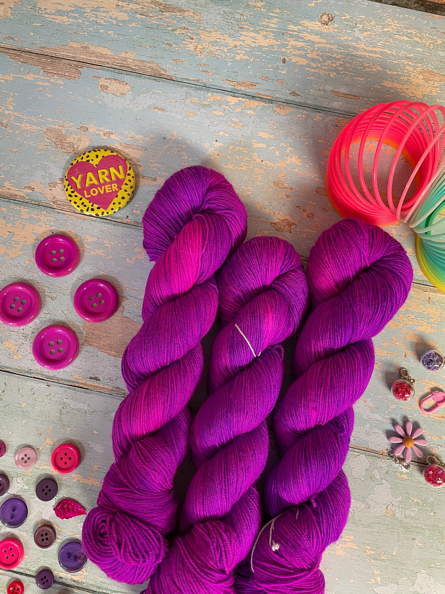 Zoomed in shot of 100g sock weight yarn in semi-solid colourway Electric Purple. This colourway is an intense purple that breaks to neon pink. Photographed on blue wooden background surrounded by props such as buttons and stitch markers.