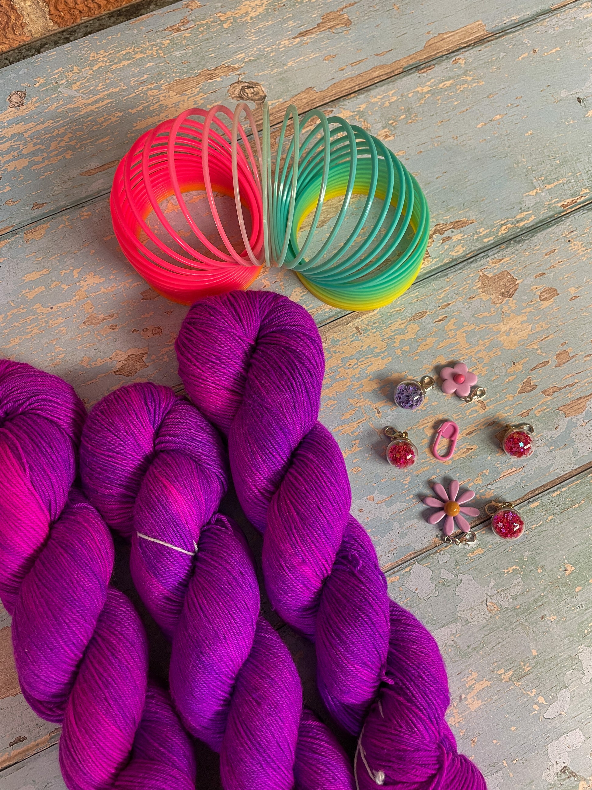 Close up shot of 100g sock weight yarn in semi-solid colourway Electric Purple. This colourway is an intense purple that breaks to neon pink. Photographed on blue wooden background surrounded by props such as buttons and stitch markers.