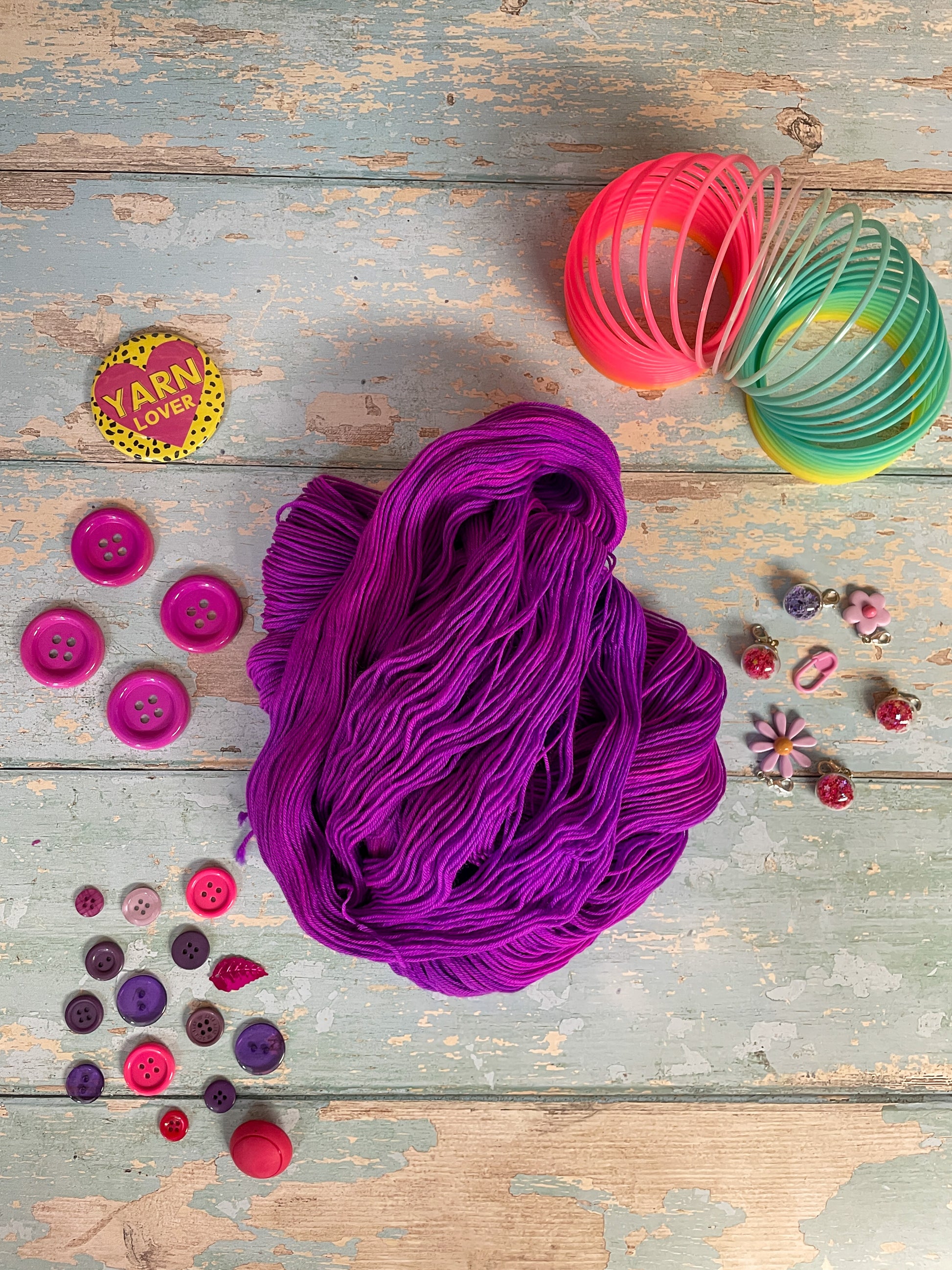 Unravelled yarn shot of 100g sock weight yarn in semi-solid colourway Electric Purple. This colourway is an intense purple that breaks to neon pink. Photographed on blue wooden background surrounded by props such as buttons and stitch markers.