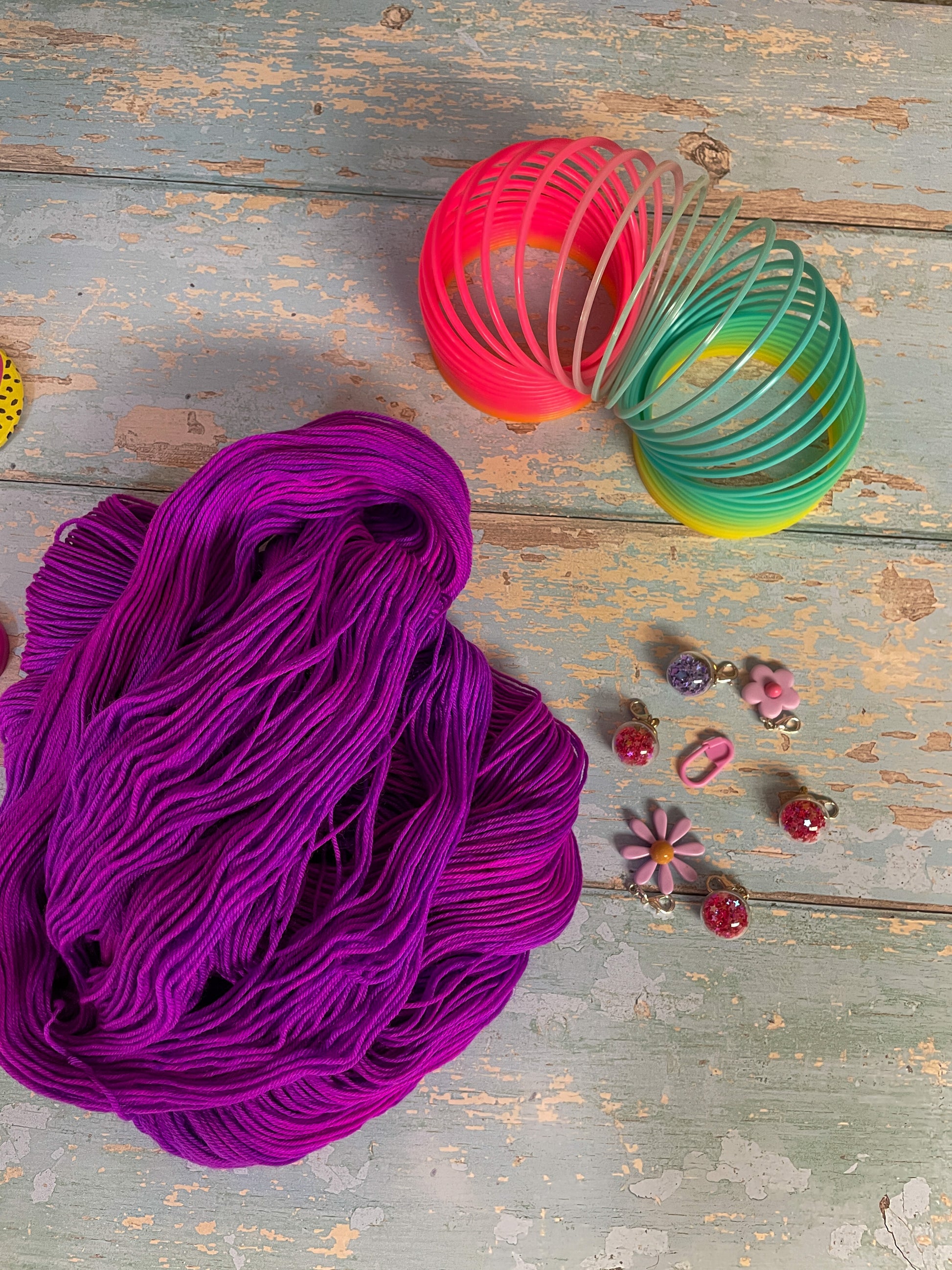 Unravelled yarn shot of 100g sock weight yarn in semi-solid colourway Electric Purple. This colourway is an intense purple that breaks to neon pink. Photographed on blue wooden background surrounded by props such as buttons and stitch markers.