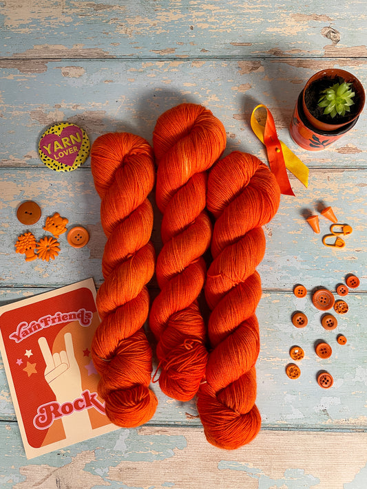 Full shot of 100g Sock skeins in semi-solid colourway Lava, an intense and bright Orange. The skeins are surrounded by props; stitch markers, buttons and Yarn Friends Rock postcard.