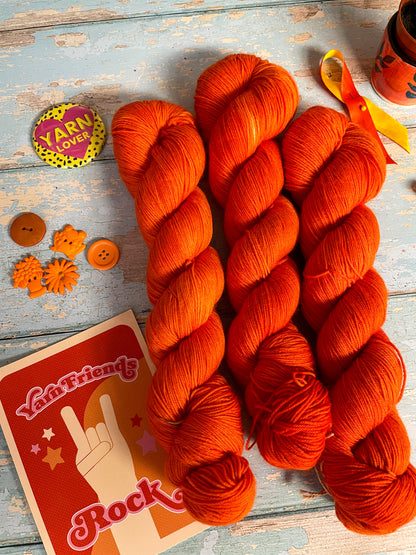 Zoomed in shot of 100g Sock skeins in semi-solid colourway Lava, an intense and bright Orange. The skeins are surrounded by props; stitch markers, buttons and Yarn Friends Rock postcard.