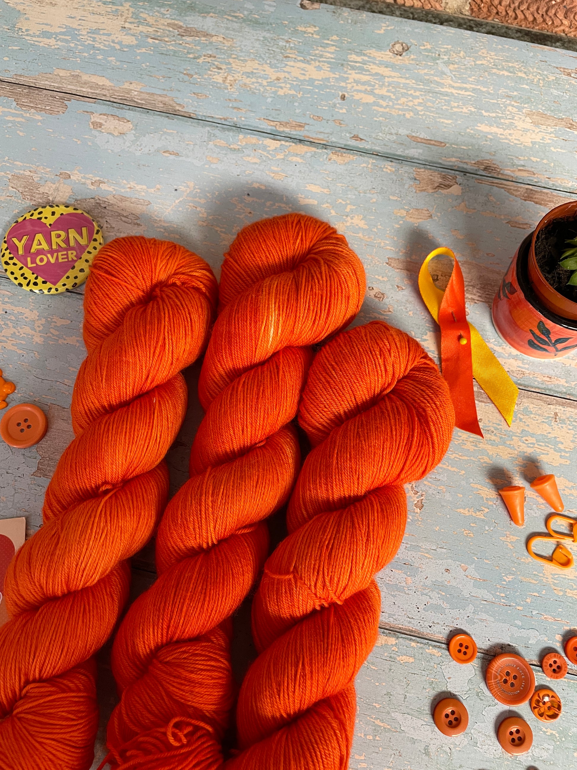 Close up of 100g Sock skeins in semi-solid colourway Lava, an intense and bright Orange. The skeins are surrounded by props; stitch markers, buttons and Yarn Lover magnet.