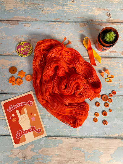 Full shot of a 100g Sock skein in semi-solid colourway Lava (untwisted), an intense and bright Orange. The untwisted skein is surrounded by props; stitch markers, buttons and Yarn Friends Rock postcard.