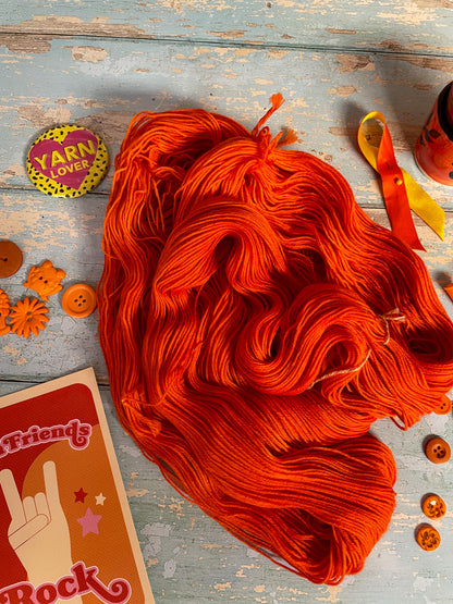 Close up shot of a 100g Sock skein in semi-solid colourway Lava (untwisted), an intense and bright Orange. The untwisted skein is surrounded by props; stitch markers, buttons and Yarn Friends Rock postcard.