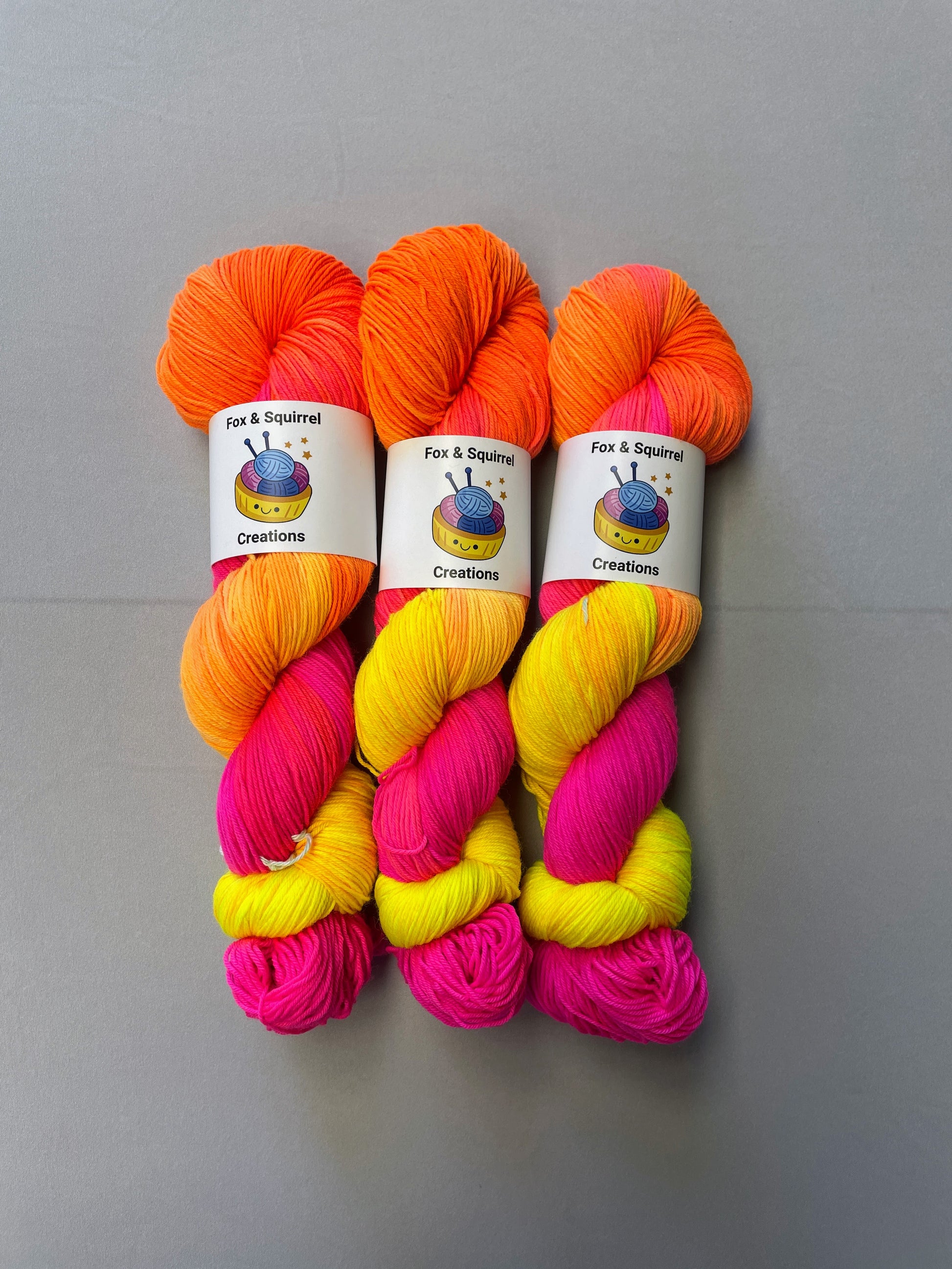 Full shot of 100g Sock weight yarn in colourway Neon Funk. This colourway has three bands of colour in Neon Yellow, Neon Pink and Neon Orange. Photographed on a light grey background. 