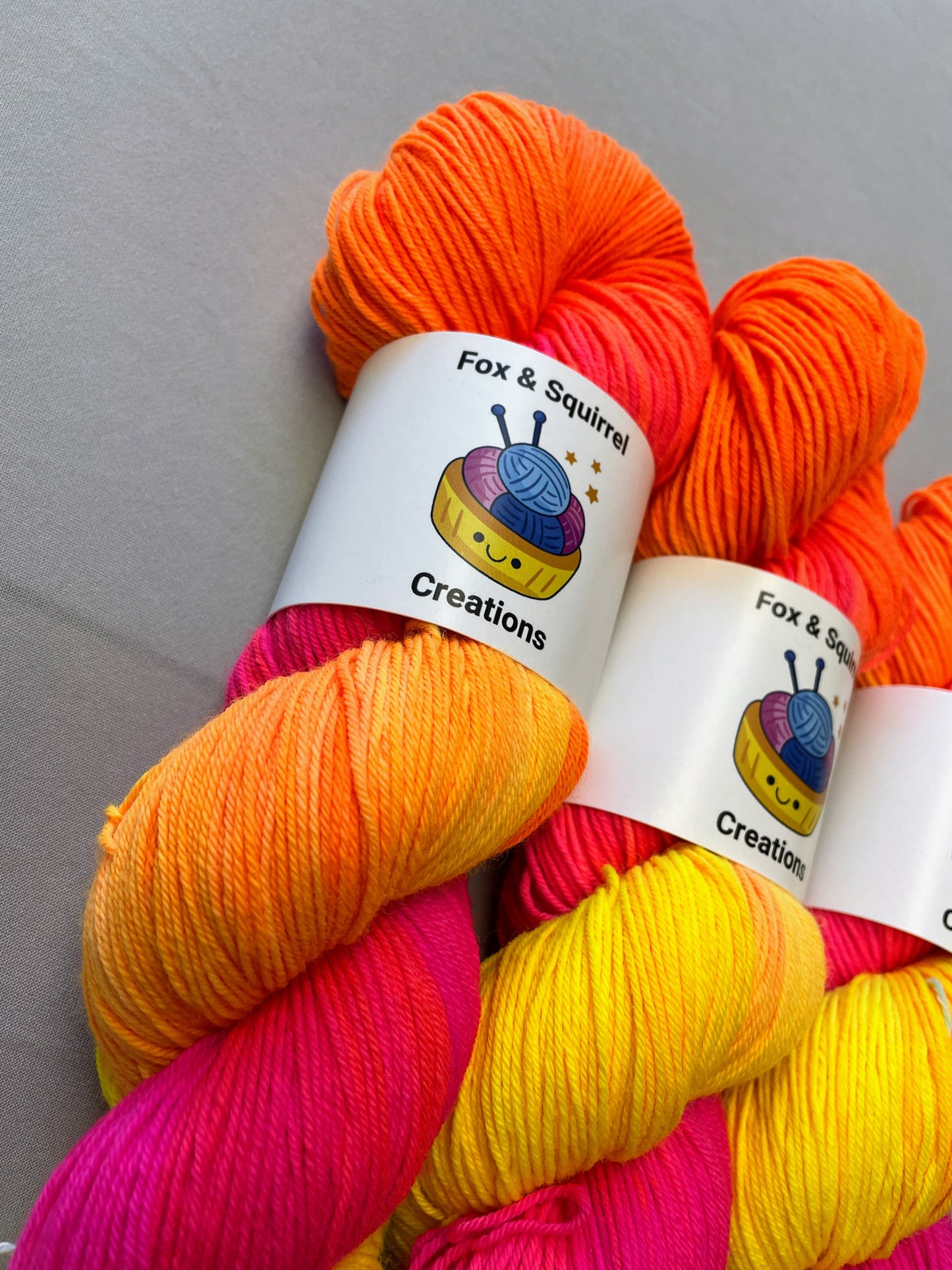 Close up shot of 100g Sock weight yarn in colourway Neon Funk. This colourway has three bands of colour in Neon Yellow, Neon Pink and Neon Orange. Photographed on a light grey background. 