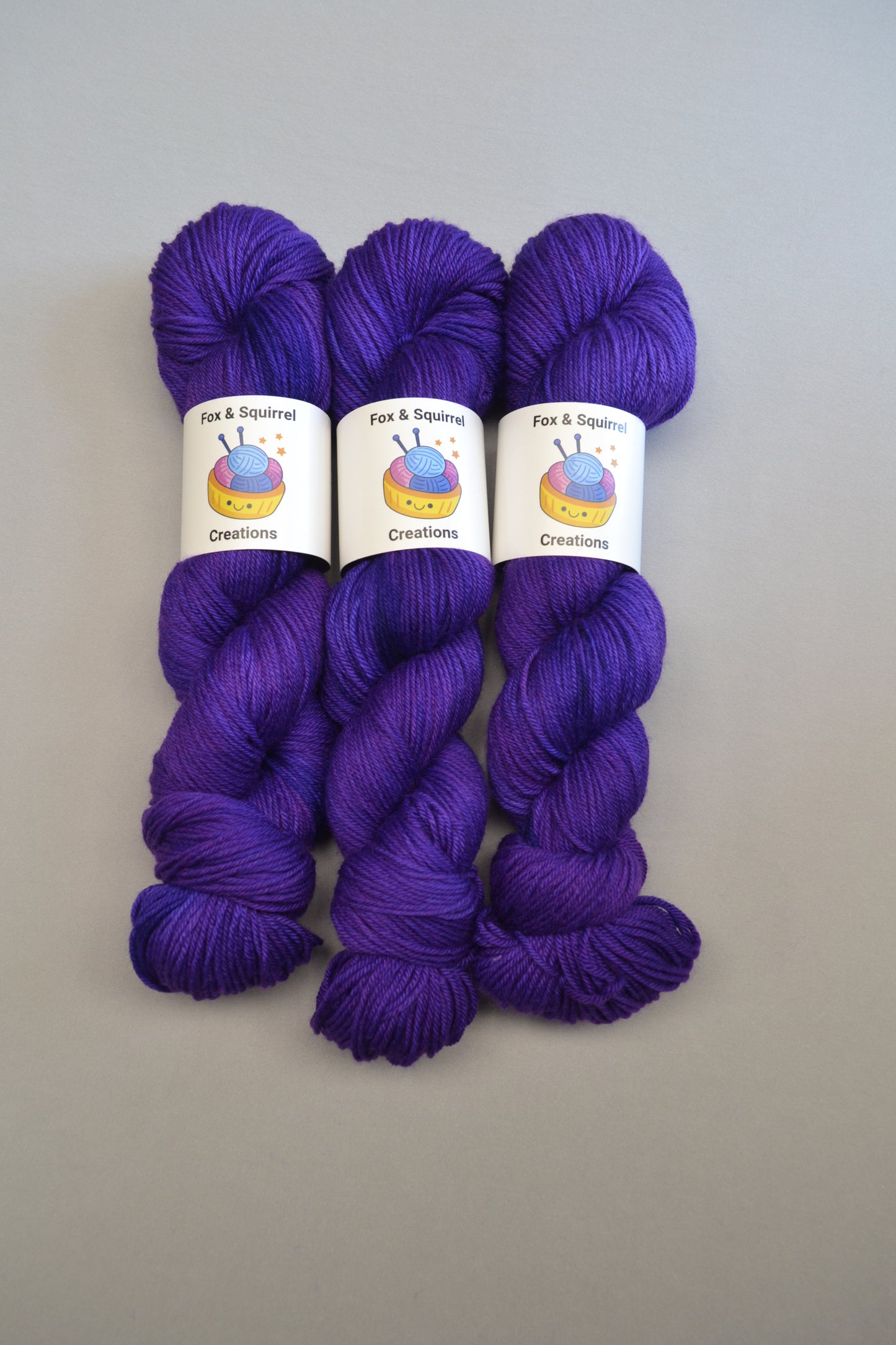 Full shot of 100g Sock weight yarn in semi-solid colourway Purple. This colourway is a deep and intense purple. Photographed on light grey background. 