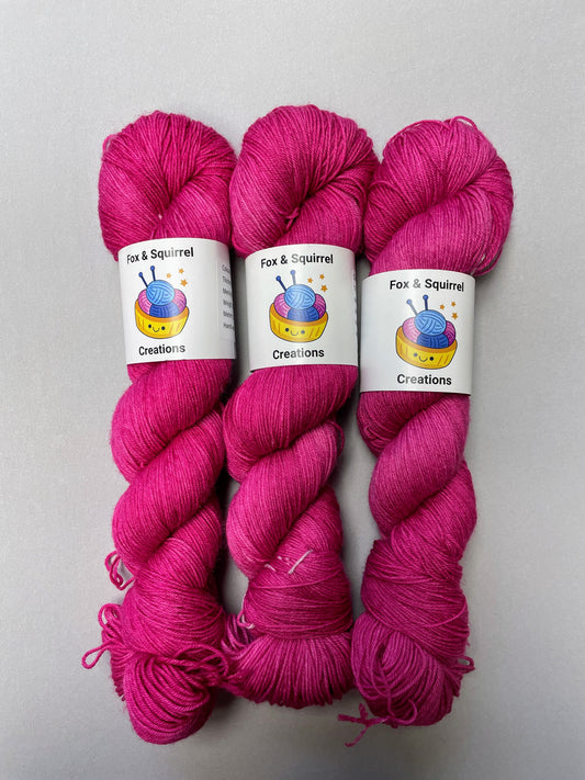 Full shot of 100g Sock weight yarn in semi-solid colourway Raspberry, an intense, deep pink. Photographed on a light grey background.