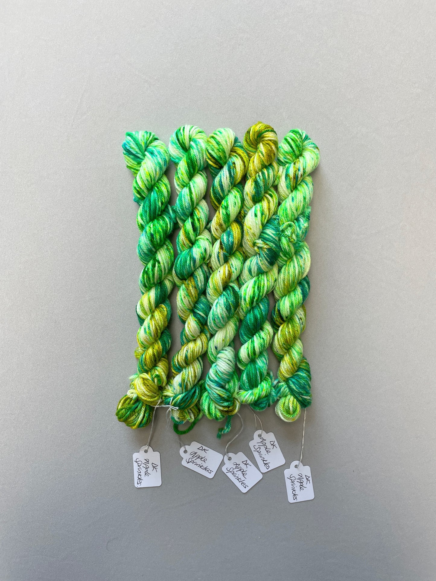 Full shot of 20g DK weight mini skeins in colourway Apple Sprinkles. Undyed yarn with layers of green speckles in shades Emerald, Olive Green and Neon Green. 