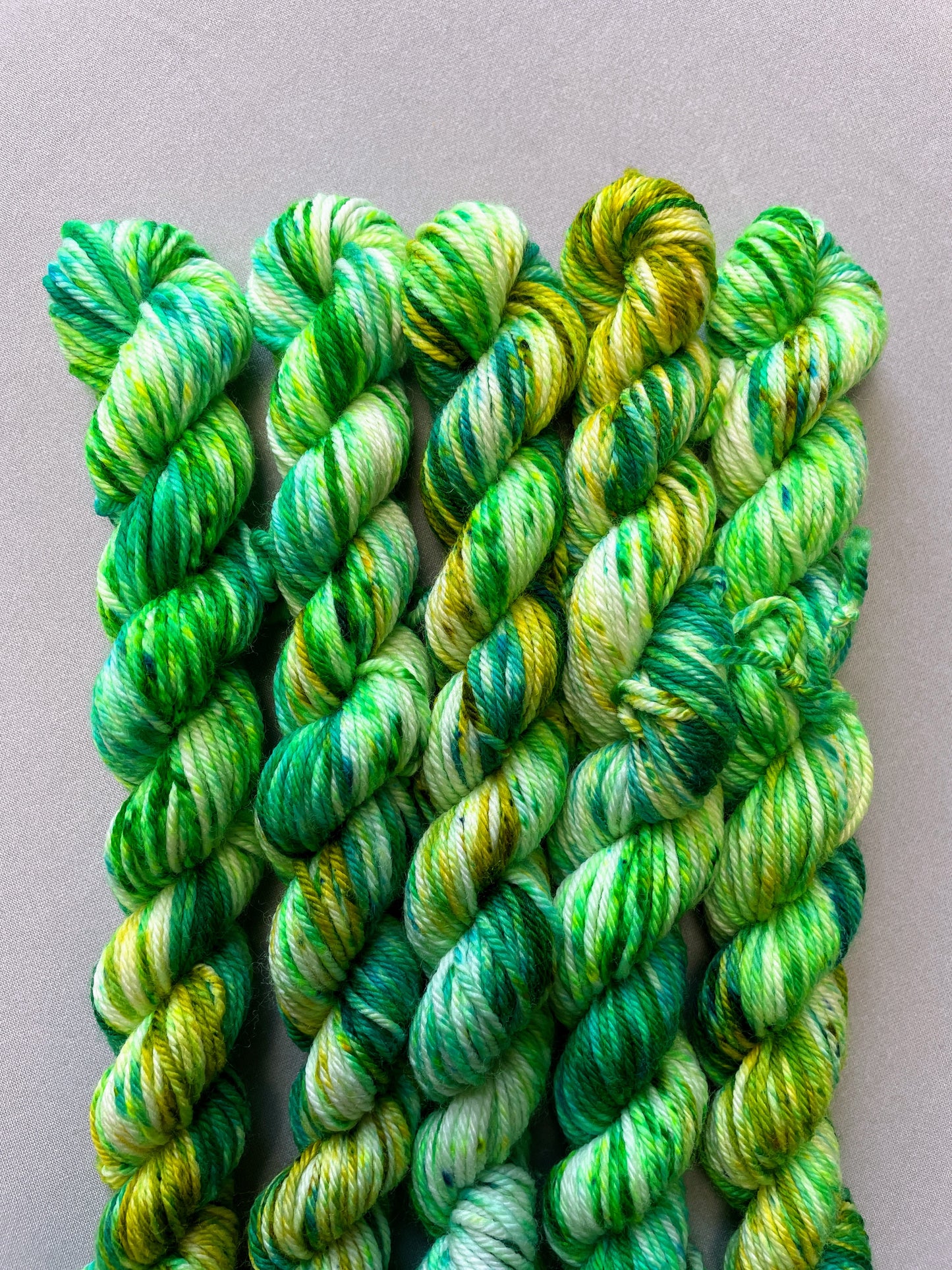 Close up shot of 20g DK weight mini skeins in colourway Apple Sprinkles. Undyed yarn with layers of green speckles in shades Emerald, Olive Green and Neon Green. 