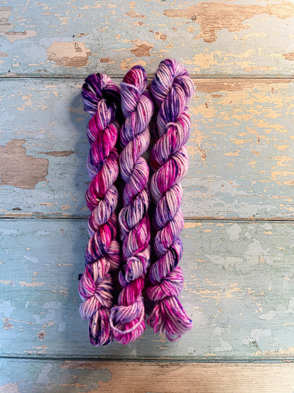 Full shot of 20g DK weight mini skeins in colourway Berry Sprinkles. Undyed yarn with layers of speckles in Pink, Neon Pink and Purple. Photographed on a blue wooden background. 