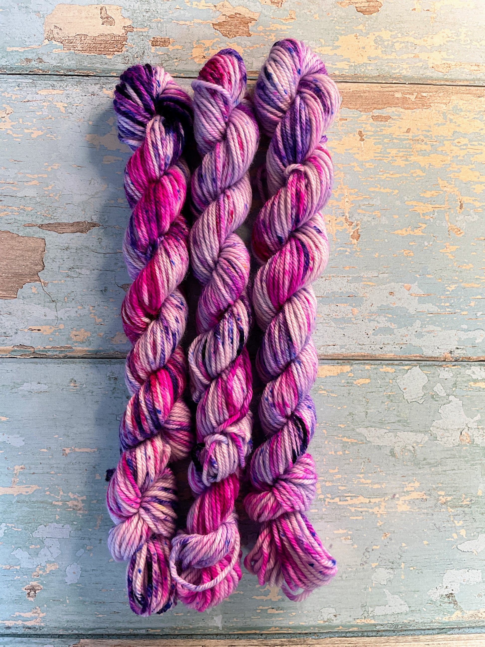 Zoomed in shot of 20g DK weight mini skeins in colourway Berry Sprinkles. Undyed yarn with layers of speckles in Pink, Neon Pink and Purple. Photographed on a blue wooden background. 