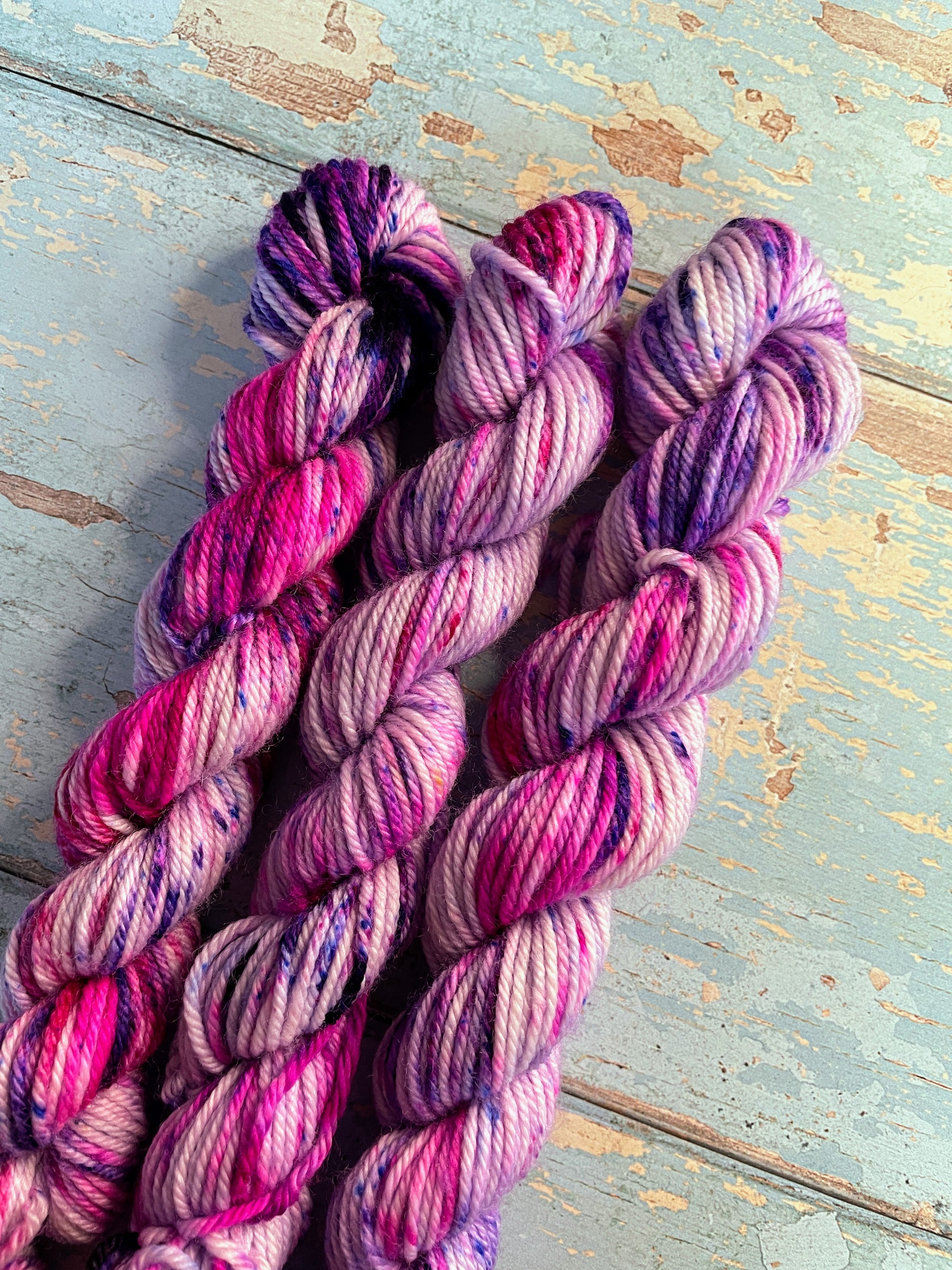 Close up shot of 20g DK weight mini skeins in colourway Berry Sprinkles. Undyed yarn with layers of speckles in Pink, Neon Pink and Purple. Photographed on a blue wooden background. 