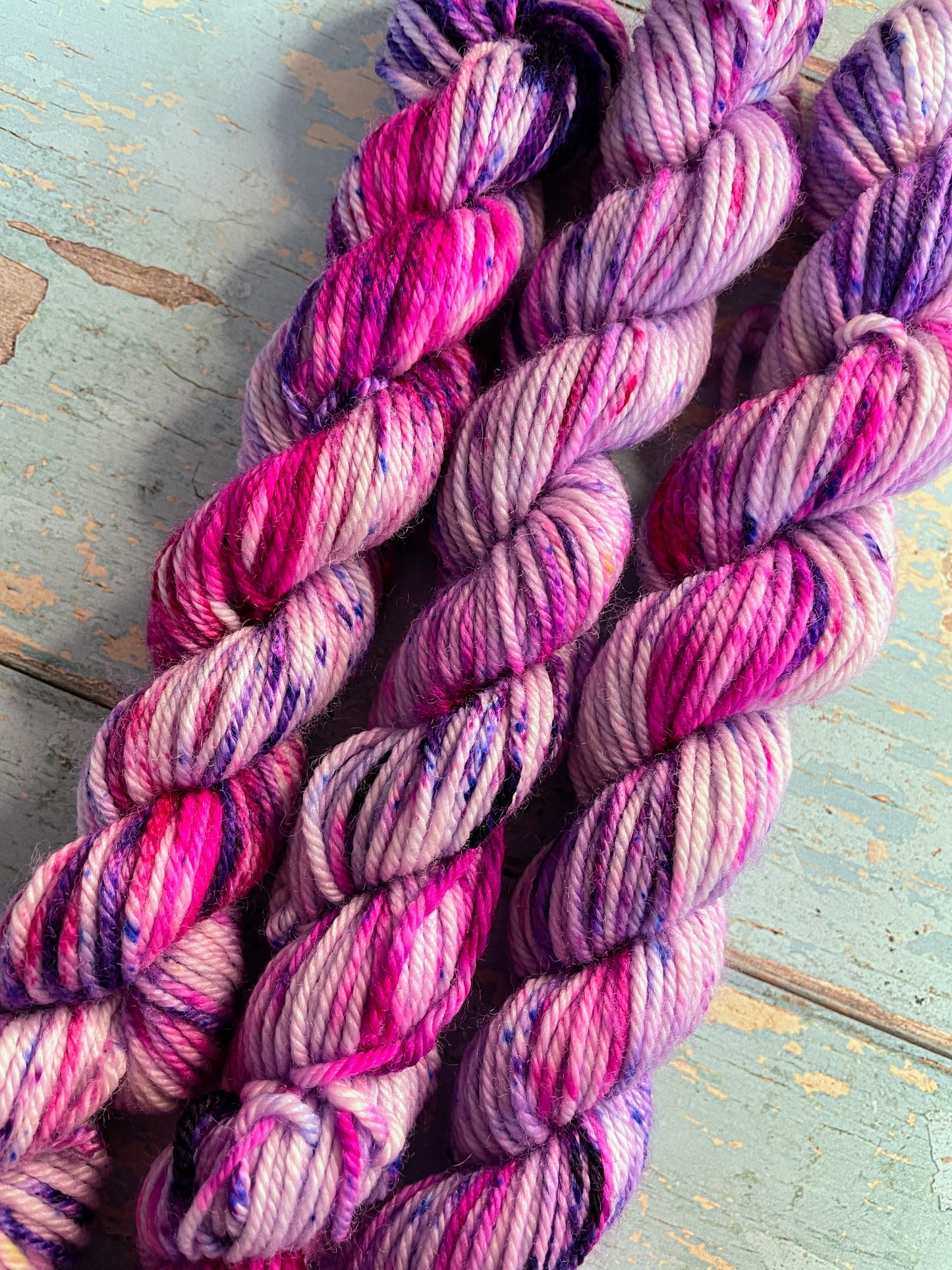 Close up shot of 20g DK weight mini skeins in colourway Berry Sprinkles. Undyed yarn with layers of speckles in Pink, Neon Pink and Purple. Photographed on a blue wooden background. 