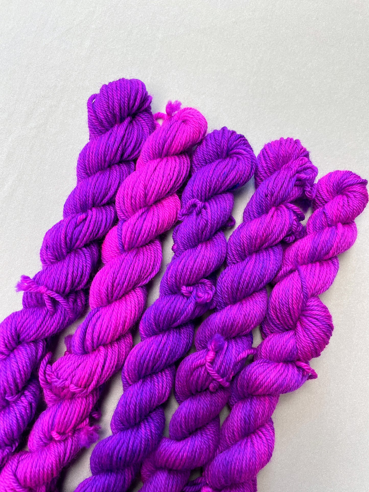 Angled shot of 20g DK weigh mini skeins in colourway Electric Purple. This colourway is an intense and bright purple that breaks into Neon Pink. Photographed on light grey background.