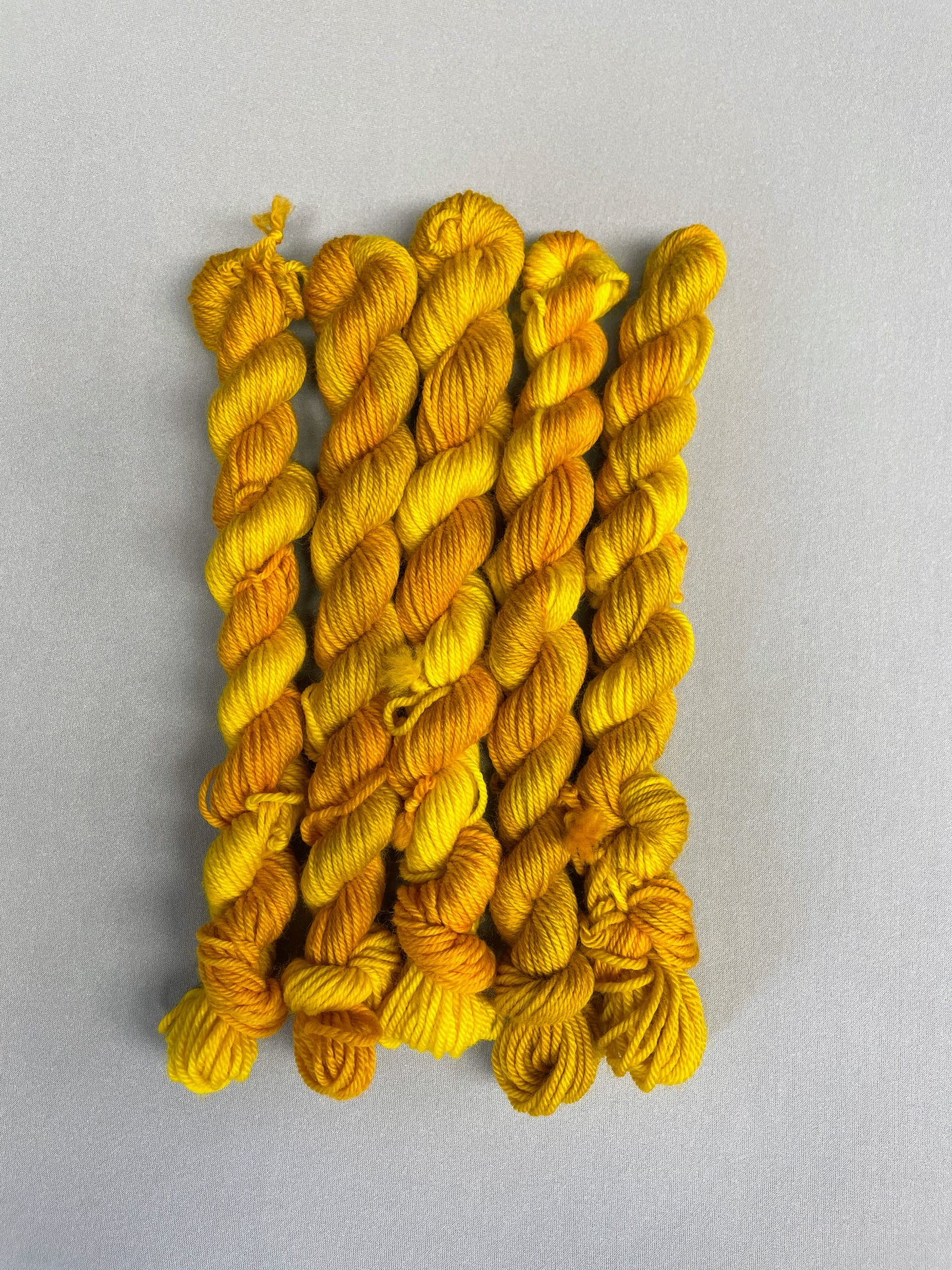 5 x 20g DK mini skeins in colourway Golden Harvest, photographed on a light grey background. Each skein is twisted and shows tonal variations of Mustard, Yellow and Golden Poppy. 