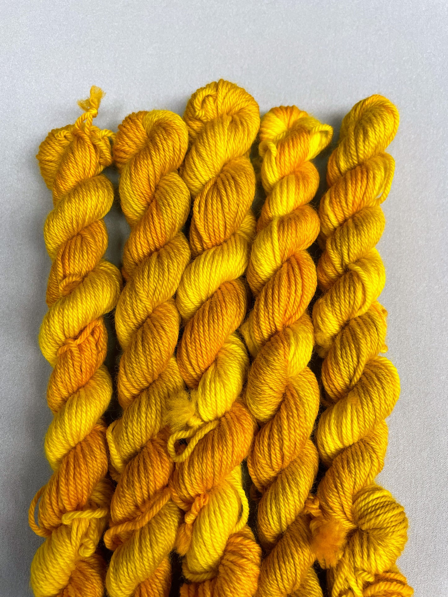 5 x 20g DK mini skeins in colourway Golden Harvest, photographed on a light grey background. Each skein is twisted and shows tonal variations of Mustard, Yellow and Golden Poppy. 