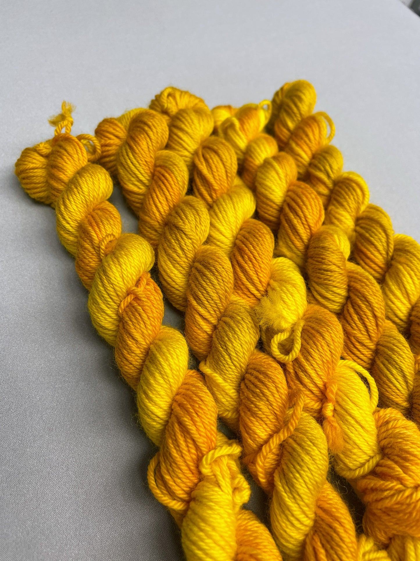 5 x 20g DK mini skeins in colourway Golden Harvest, photographed on a light grey background. Each skein is twisted and shows tonal variations of Mustard, Yellow and Golden Poppy. 