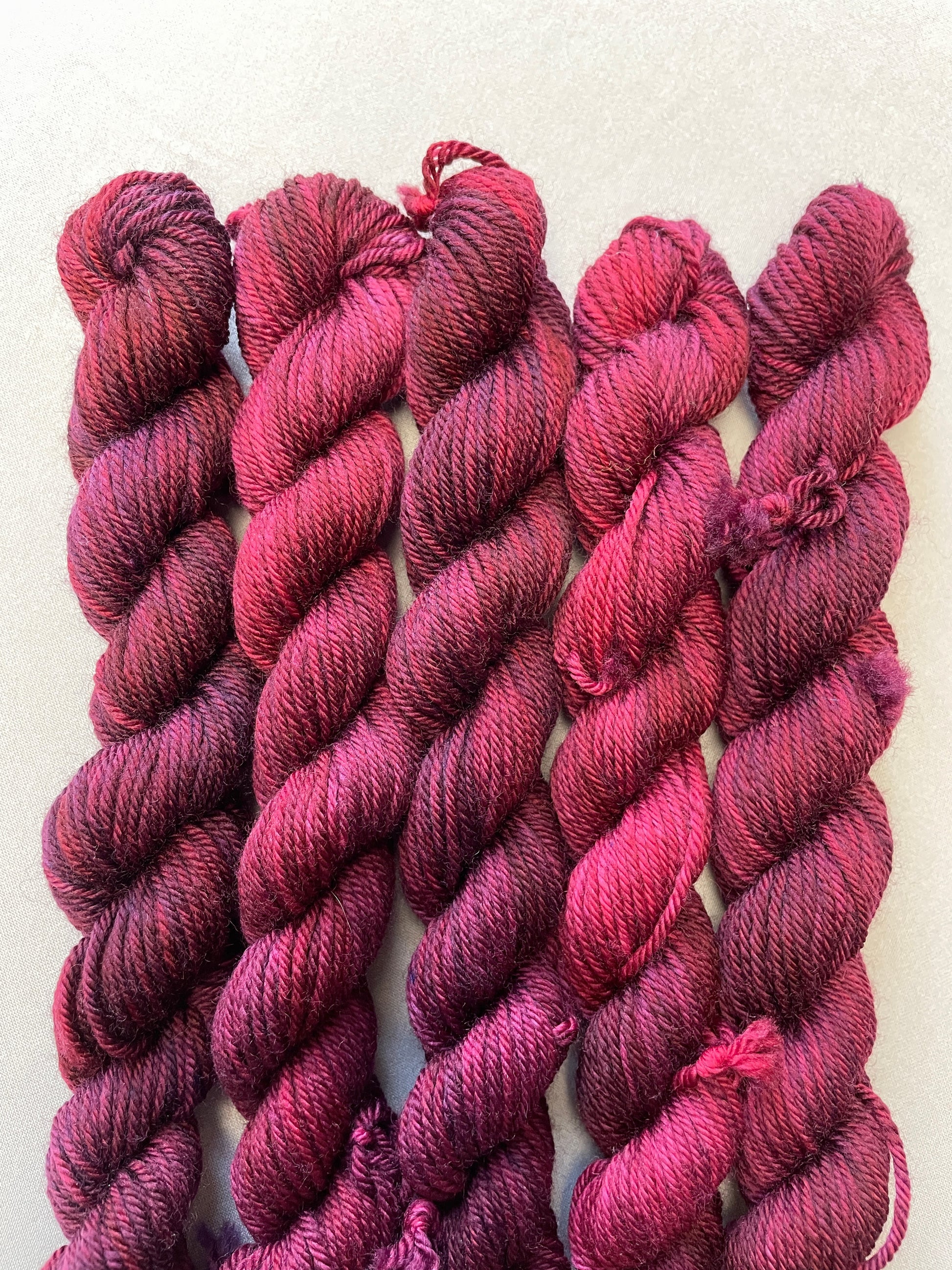 Close up shot of 20g DK weight mini skeins in semi-solid colourway Plum an intense, deep Plum/Burgundy. Photographed on a light grey background.