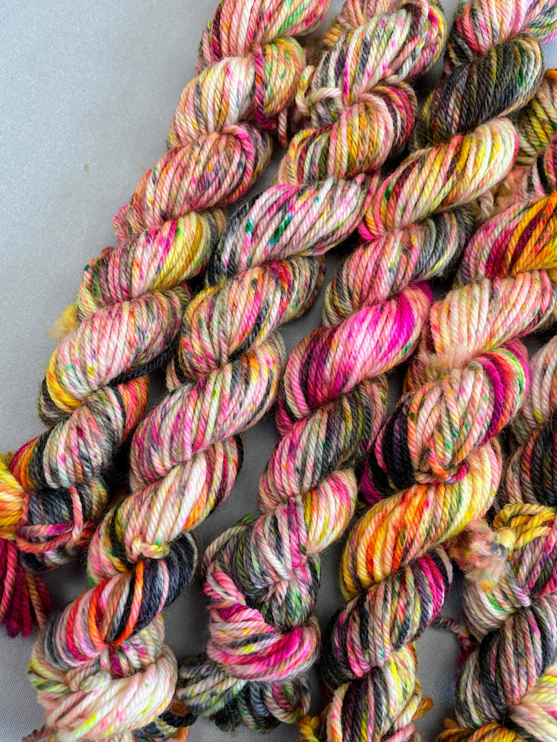 Close up in shot of 20g DK mini skeins in colourway Punk Rocker. An undyed yarn base with layers of Neon sprinkles in Yellow, Pink, Green and Black. This colourway glows under U.V. light. 