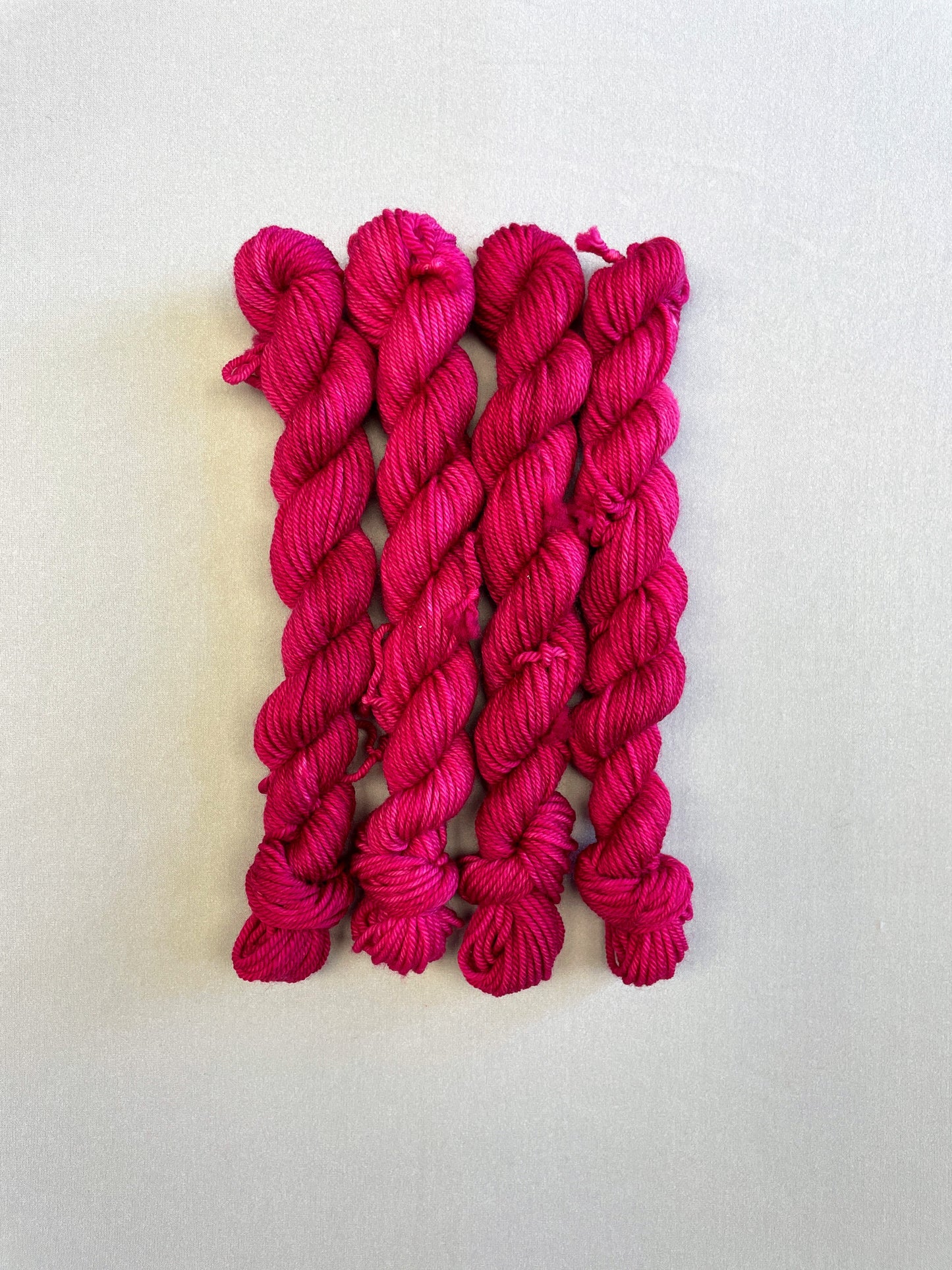 Full shot of 20g DK weight mini skeins in semi-solid colourway Raspberry, an intense, deep pink. Photographed on a light grey background.