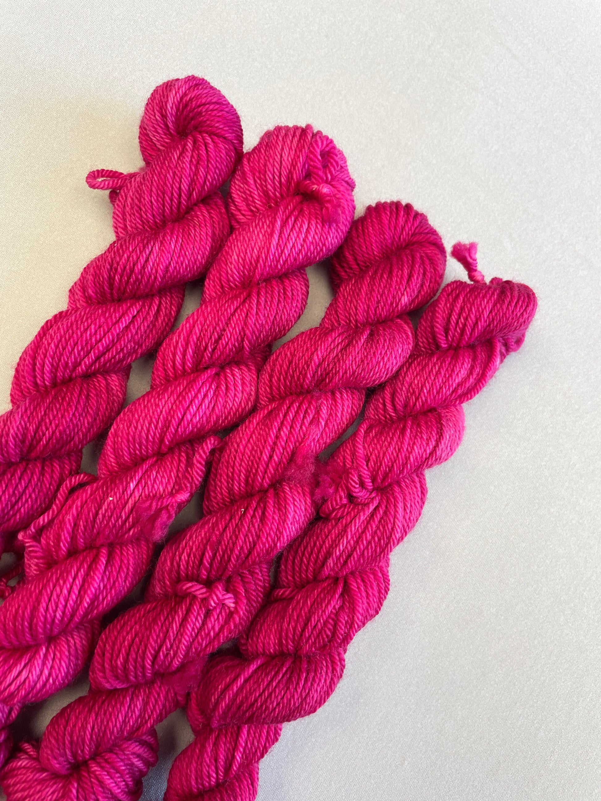 Close up shot of 20g DK weight mini skeins in semi-solid colourway Raspberry, an intense, deep pink. Photographed on a light grey background.