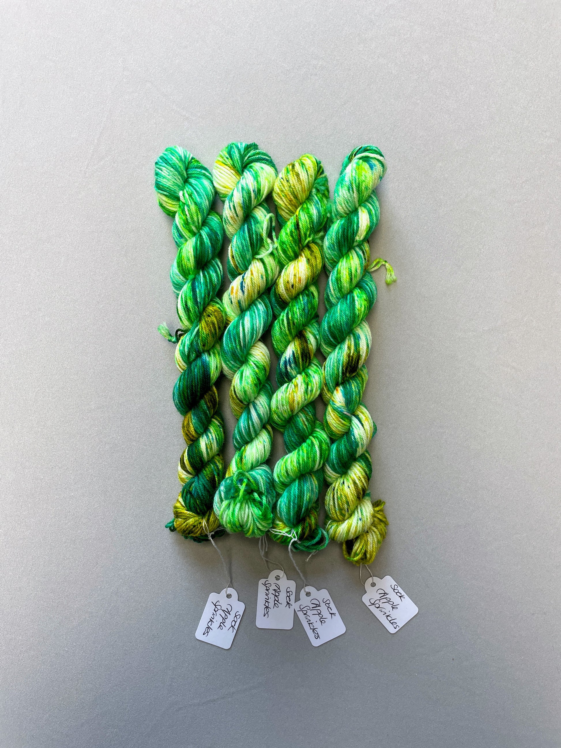 Sock weight 20g mini skein in colourway Apple Sprinkles. Undyed yarn base with layers of green speckles in shades, Emerald, Olive Green and Neon Green. 