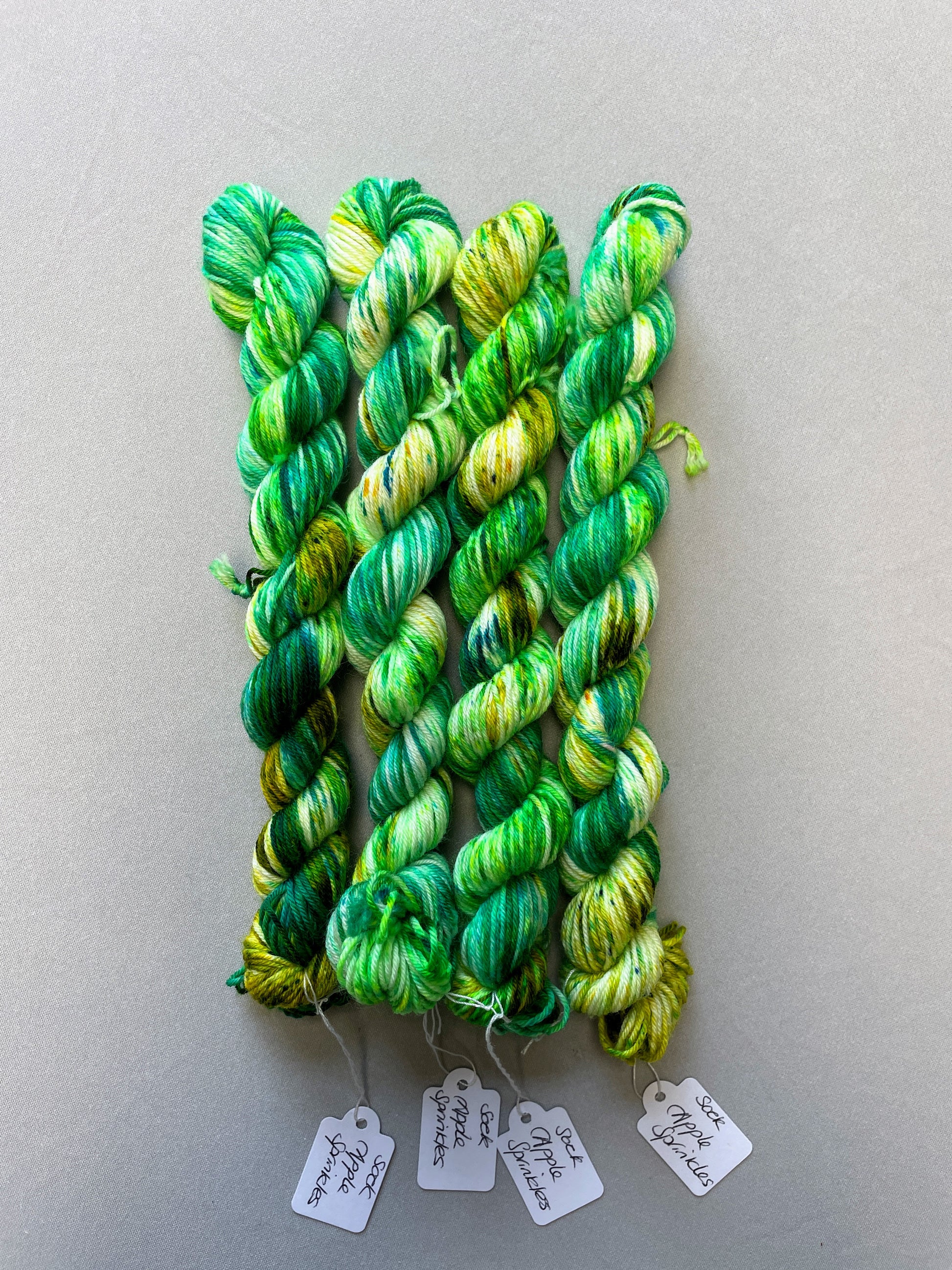 Full shot of 20g sock weight mini skein in colourway Apple Sprinkles. Undyed yarn with layers of green speckles in random placement in shades Emerald, Olive Green and Neon Green. 