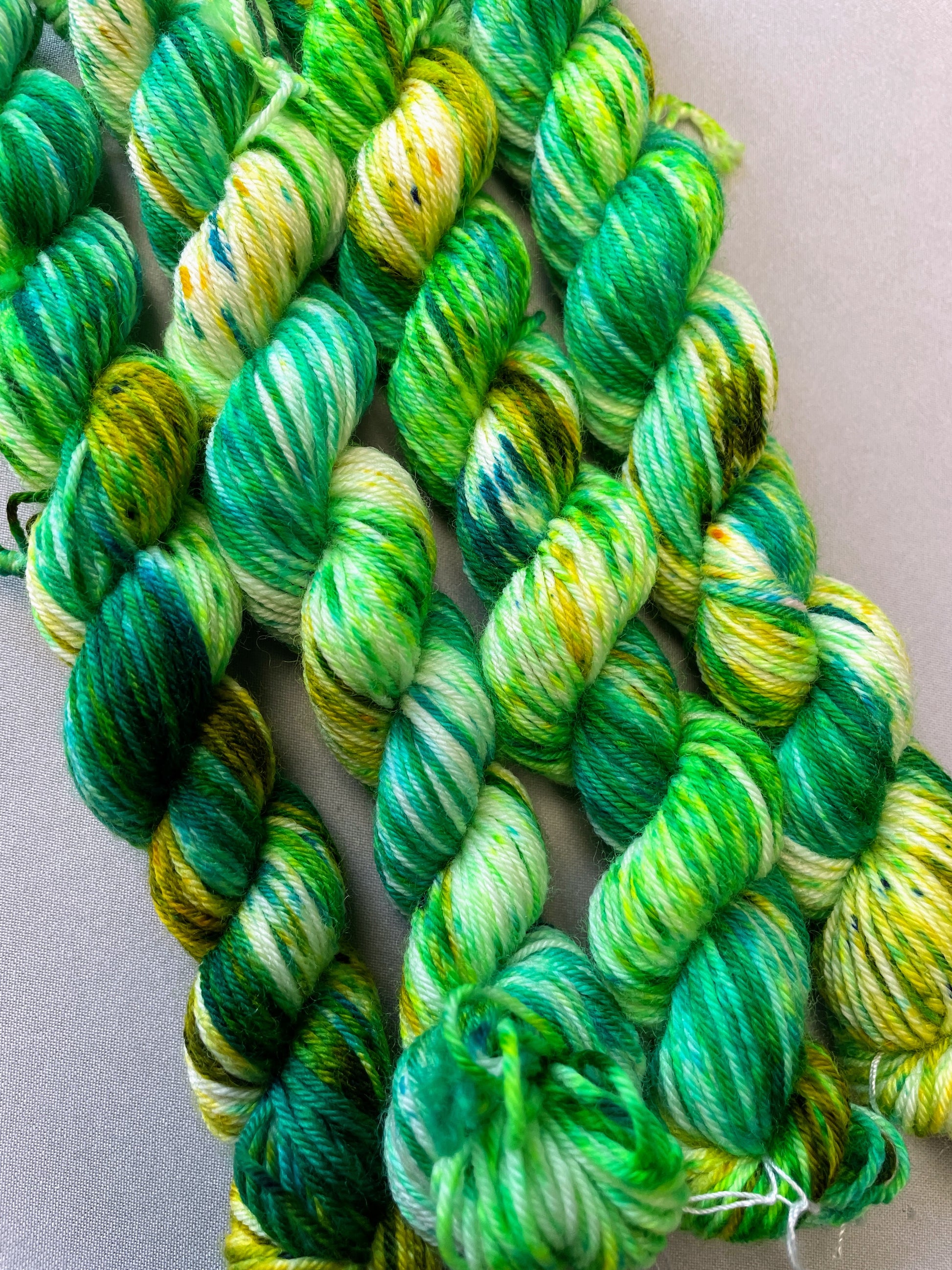 Close up shot of 20g sock weight Mini Skein in colourway Apple Sprinkles. Undyed yarn base with layers of green sprinkles in shades Emerald, Olive Green and Neon Green. 