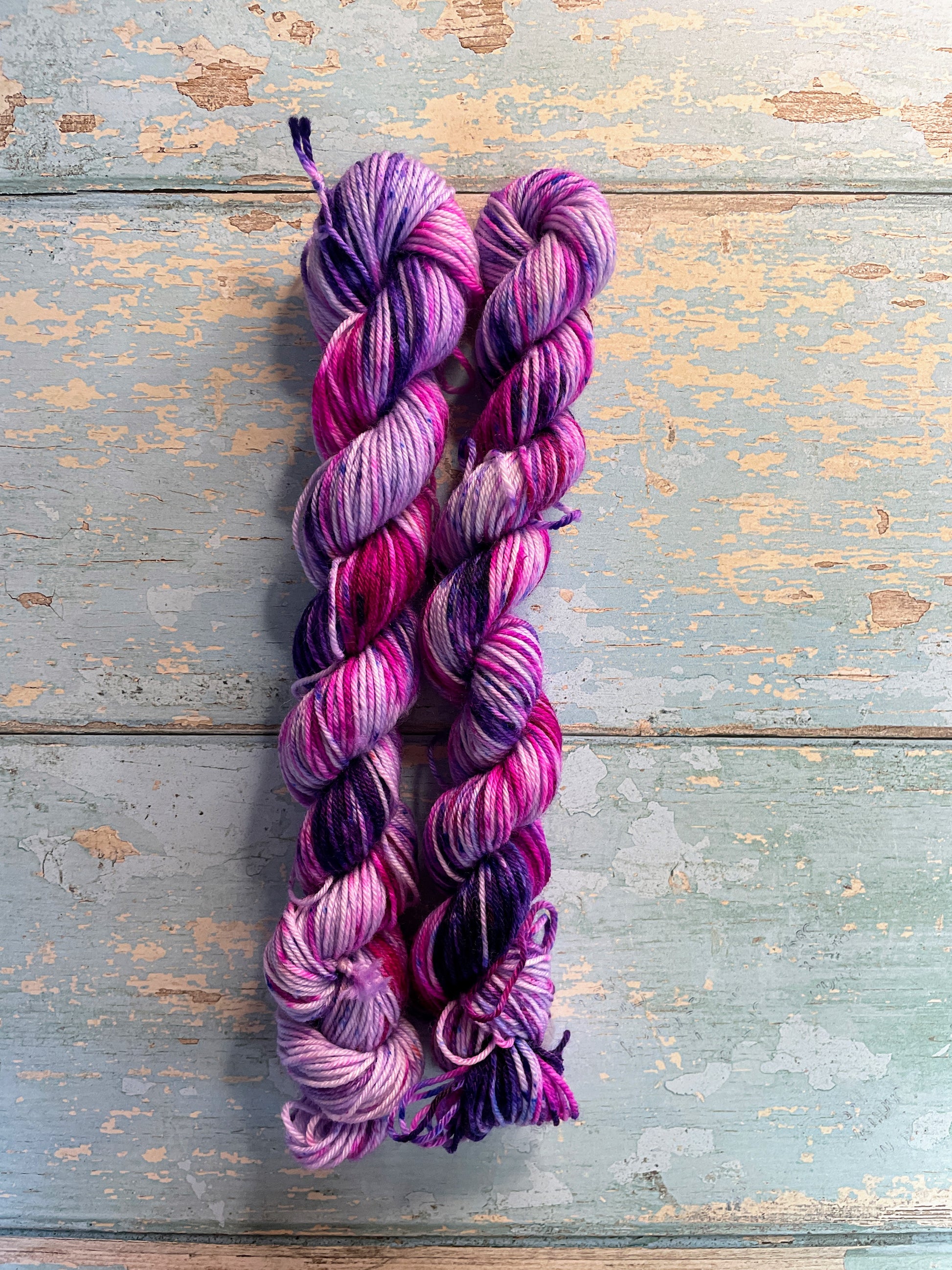 Full shot of 20g sock weight mini skeins in colourway Berry Sprinkles. Undyed yarn with layers of speckles in Pink, Neon Pink and Purple. Photographed on a blue wooden background. 