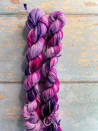 Zoomed in shot of 20g sock weight mini skeins in colourway Berry Sprinkles. Undyed yarn with layers of speckles in Pink, Neon Pink and Purple. Photographed on a blue wooden background. 