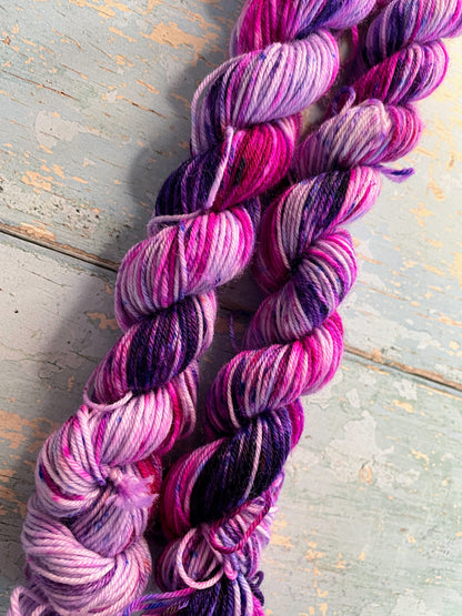 Close up shot of 20g sock weight mini skeins in colourway Berry Sprinkles. Undyed yarn with layers of speckles in Pink, Neon Pink and Purple. Photographed on a blue wooden background. 