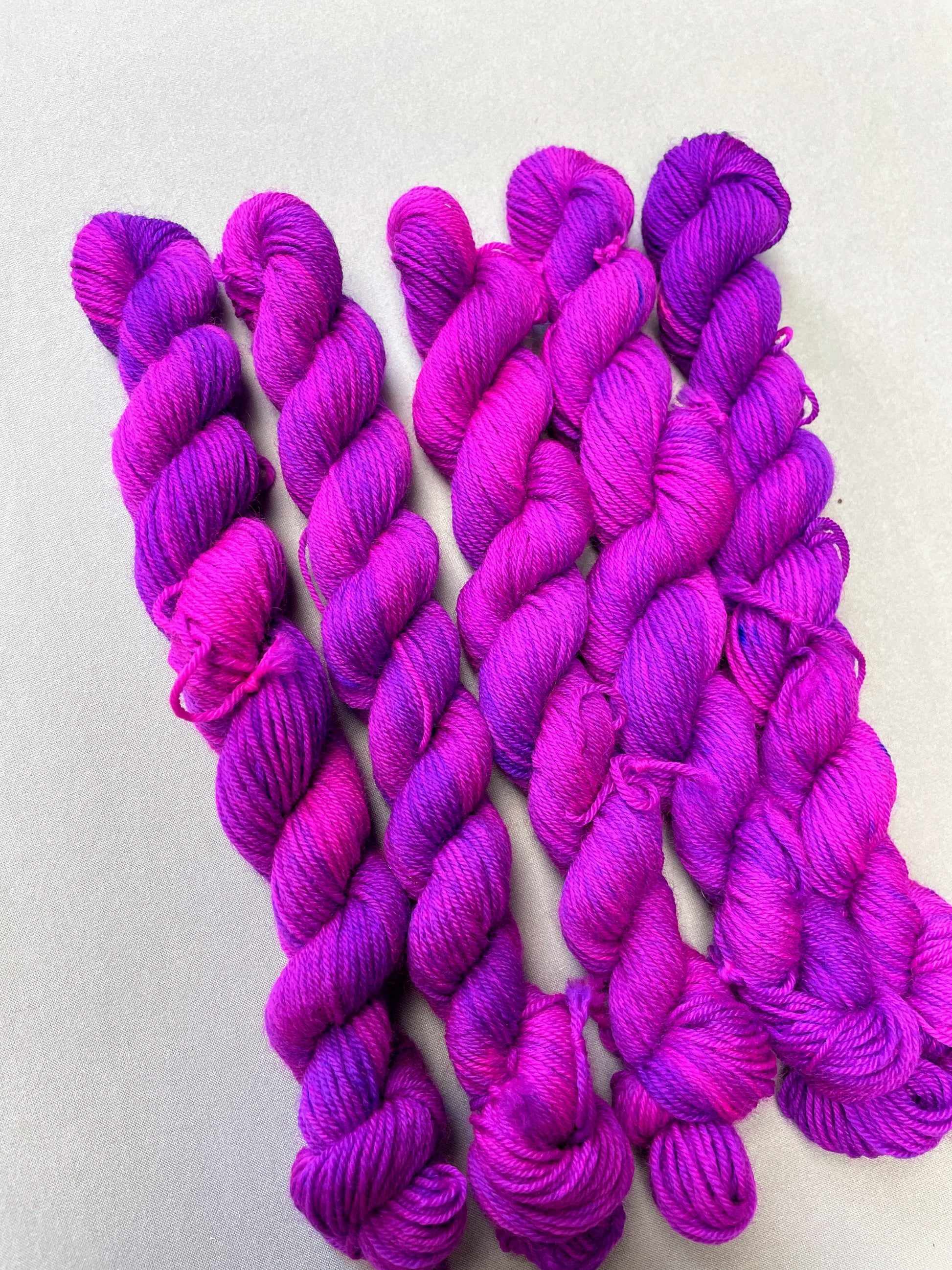 Angled shot of 20g Sock weigh mini skeins in colourway Electric Purple. This colourway is an intense and bright purple that breaks into Neon Pink. Photographed on light grey background.