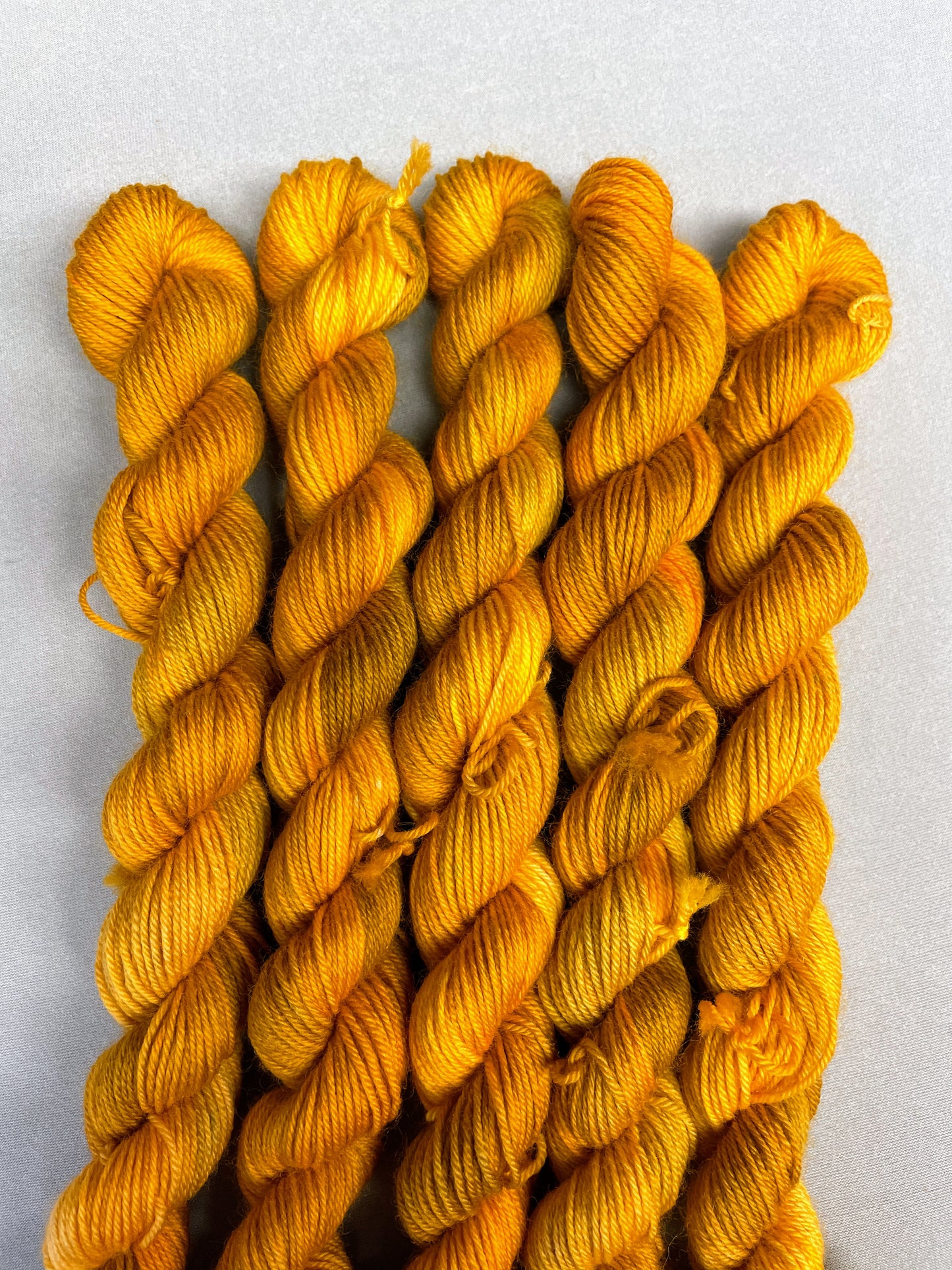 Hand-dyed Yarn 
5 x 20g, twisted, Sock mini skeins in colourway Hay Fields, photographed on a light grey background. This colourway is variegated with golden tones of Mustard, Golden Poppy and Saffron.